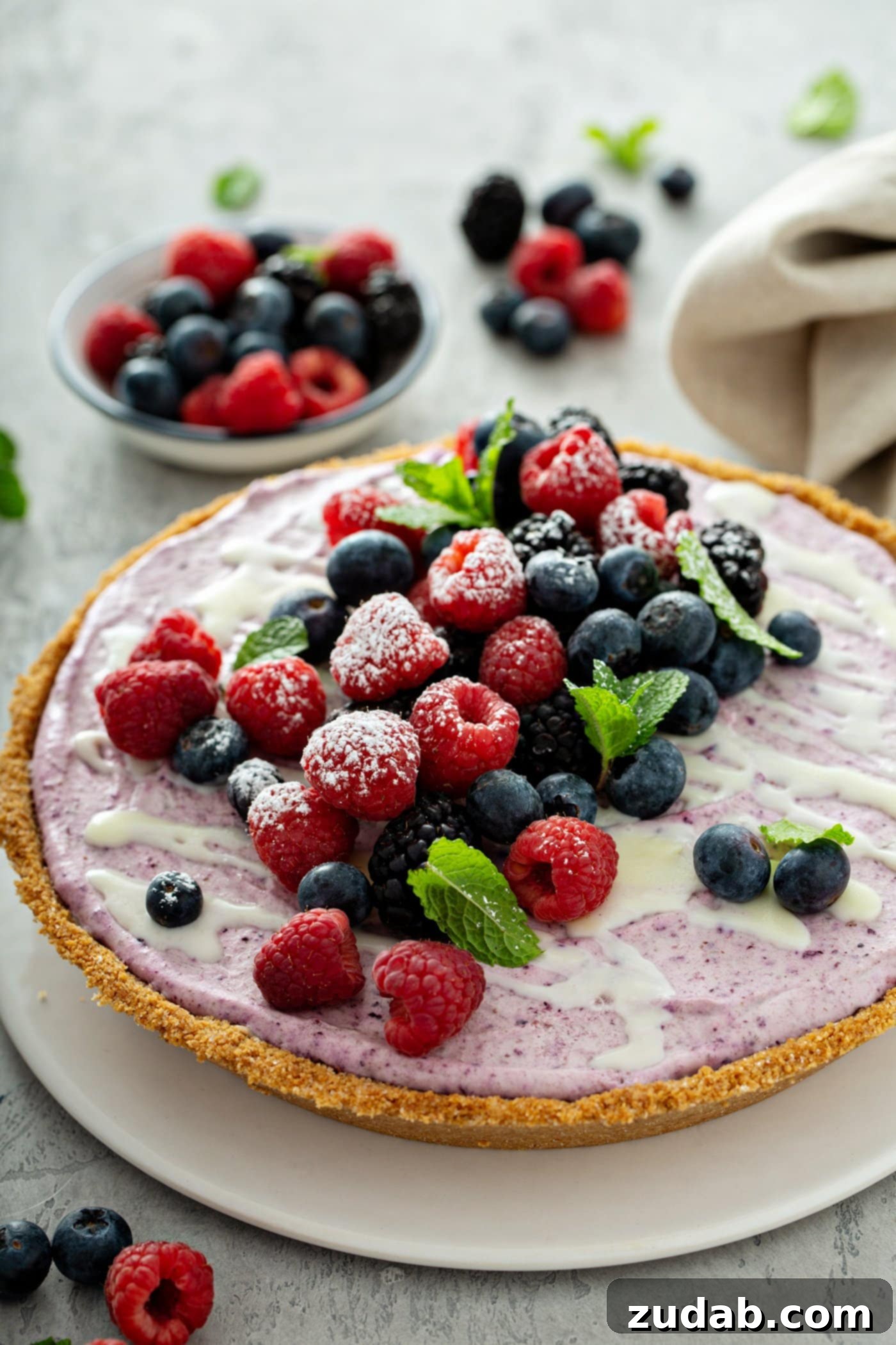 White Chocolate Berry Icebox Pie 10 A whole white chocolate berry pie. The pie is topped with whole strawberries, raspberries, blue berries, blackberries, and mint leaves. There is a bowl of mixed berries in the background, and berries and a napkin on the table.