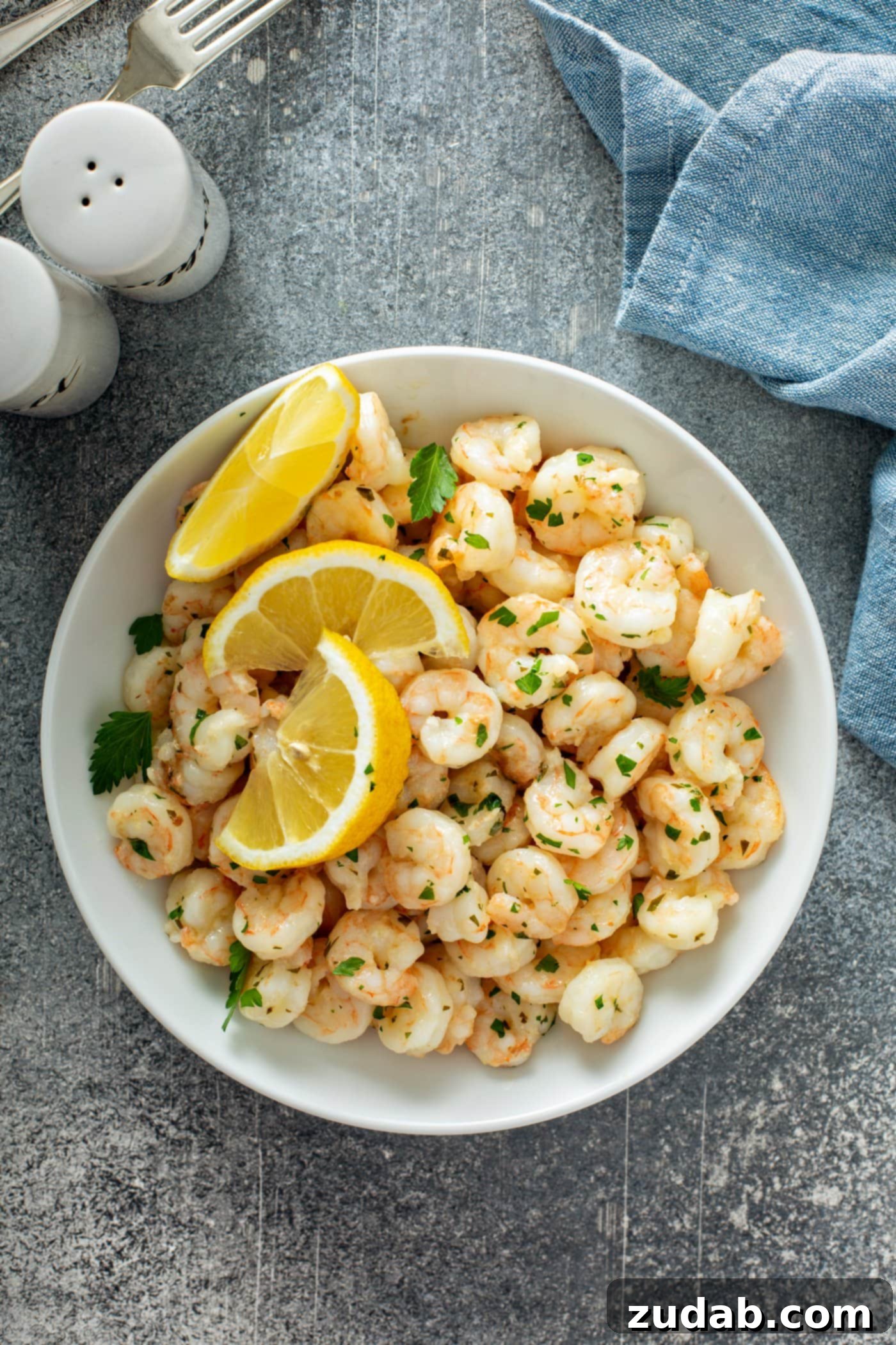 More Quick Meal Ideas A charming serving plate showcases lemon garlic shrimp, artfully arranged with three bright lemon wedges resting on top. A scattering of fresh parsley adds a vibrant touch. In the soft background, a neatly folded napkin and classic salt and pepper shakers suggest an inviting table setting.