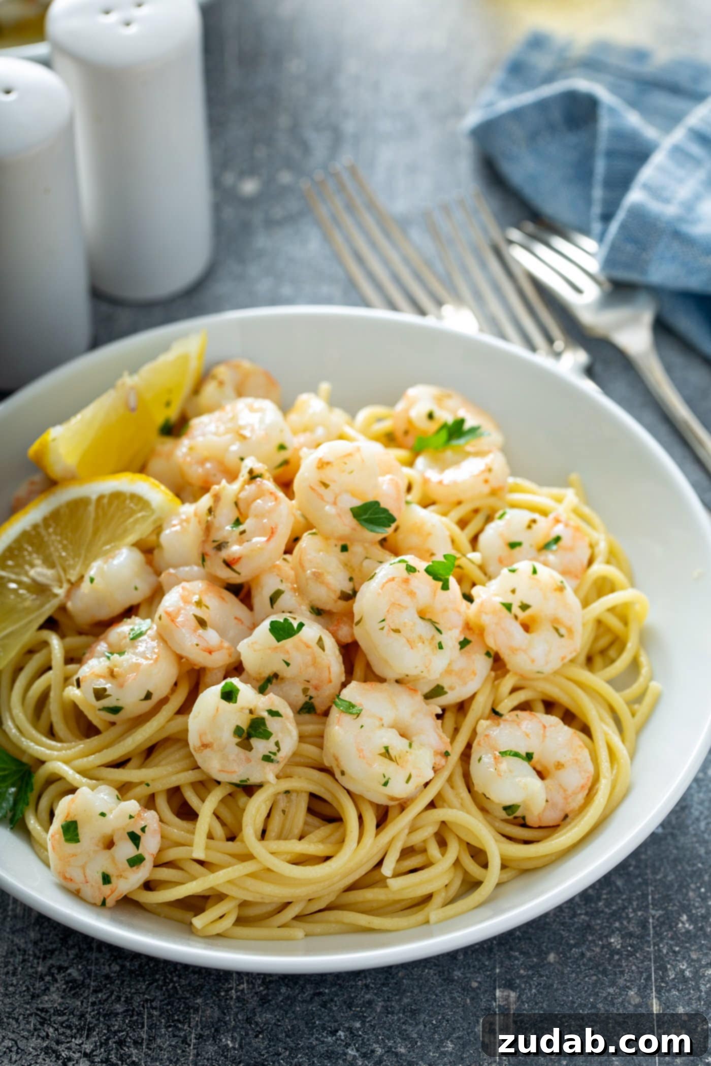 Shrimp Storage Guidelines A delightful serving dish of lemon garlic shrimp gracefully presented over a bed of pasta and adorned with fresh parsley. Two bright lemon wedges rest enticingly on the plate beside the shrimp. A charming table setting includes three forks, a classic salt and pepper shaker, and a neatly folded napkin, all ready for a memorable and quick seafood meal.