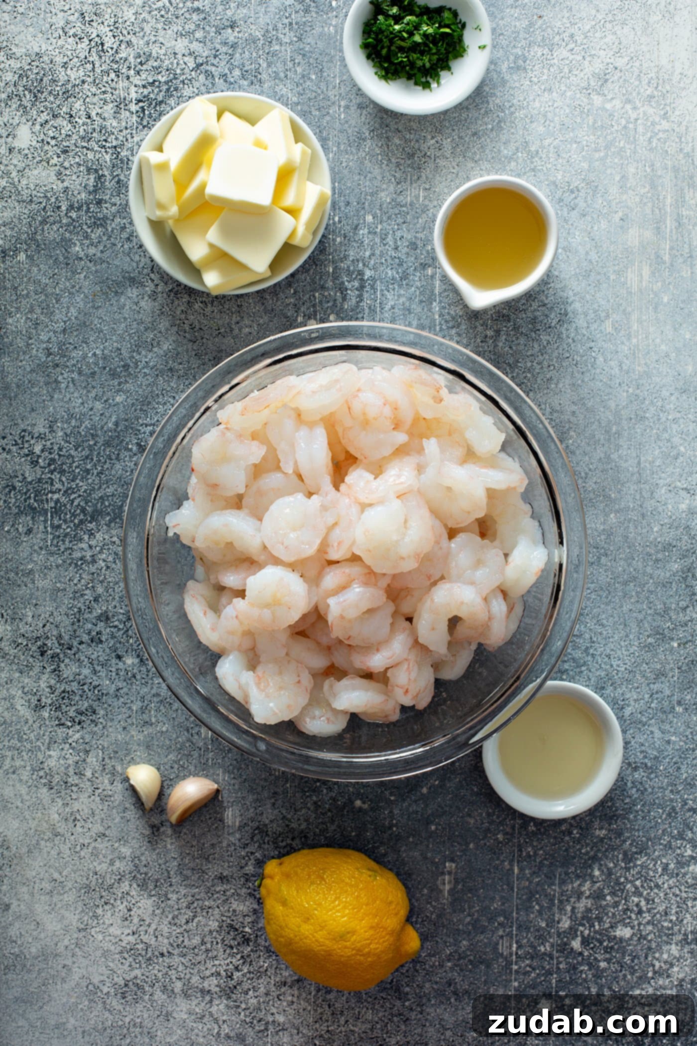 Lemon Garlic Shrimp Ingredients A glass bowl brimmed with fresh, raw shrimp. It's surrounded by small, pristine white ramekins holding individual portions of softened butter, clear chicken broth, and freshly squeezed lemon juice. Adjacent on the rustic wooden table, two robust cloves of garlic and a whole, bright yellow lemon await their turn, highlighting the fresh ingredients needed for this lemon garlic shrimp recipe.