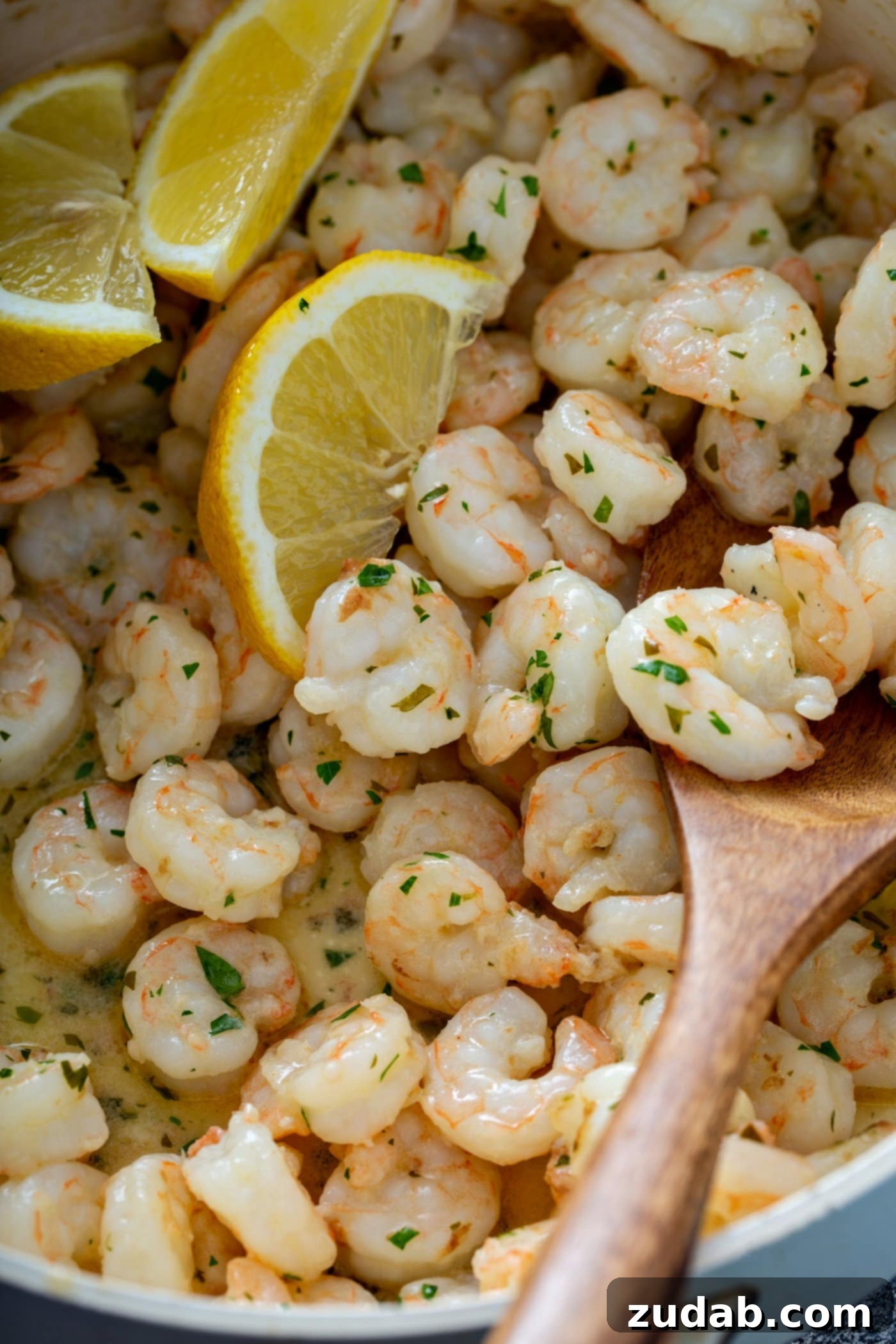 Lemon Garlic Shrimp Sauce A tantalizing close-up of lemon garlic shrimp generously coated in its luscious sauce, being scooped with a wooden spoon. Three vibrant lemon wedges are nestled among the shrimp, while fresh parsley leaves are sprinkled abundantly over the entire dish, creating an inviting aroma and visual appeal.