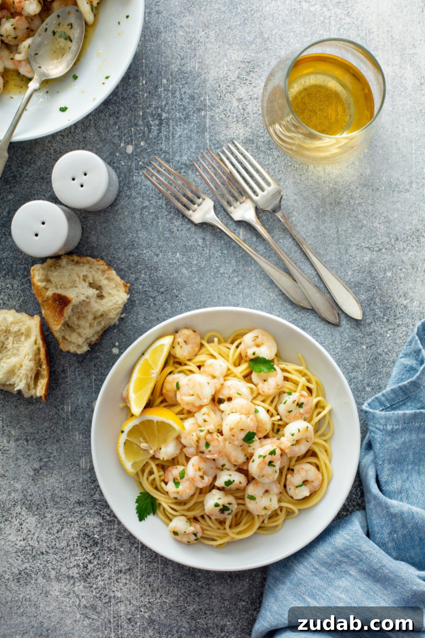 Lemon Garlic Shrimp with Pasta A beautifully presented serving dish of lemon garlic shrimp resting on a bed of pasta. Bright lemon wedges adorn the edge of the plate, and the shrimp are exquisitely sprinkled with fresh parsley. In the background, an olive oil container, three forks, salt and pepper shakers, and two pieces of rustic bread complete the inviting table setting, hinting at a complete and delicious meal.