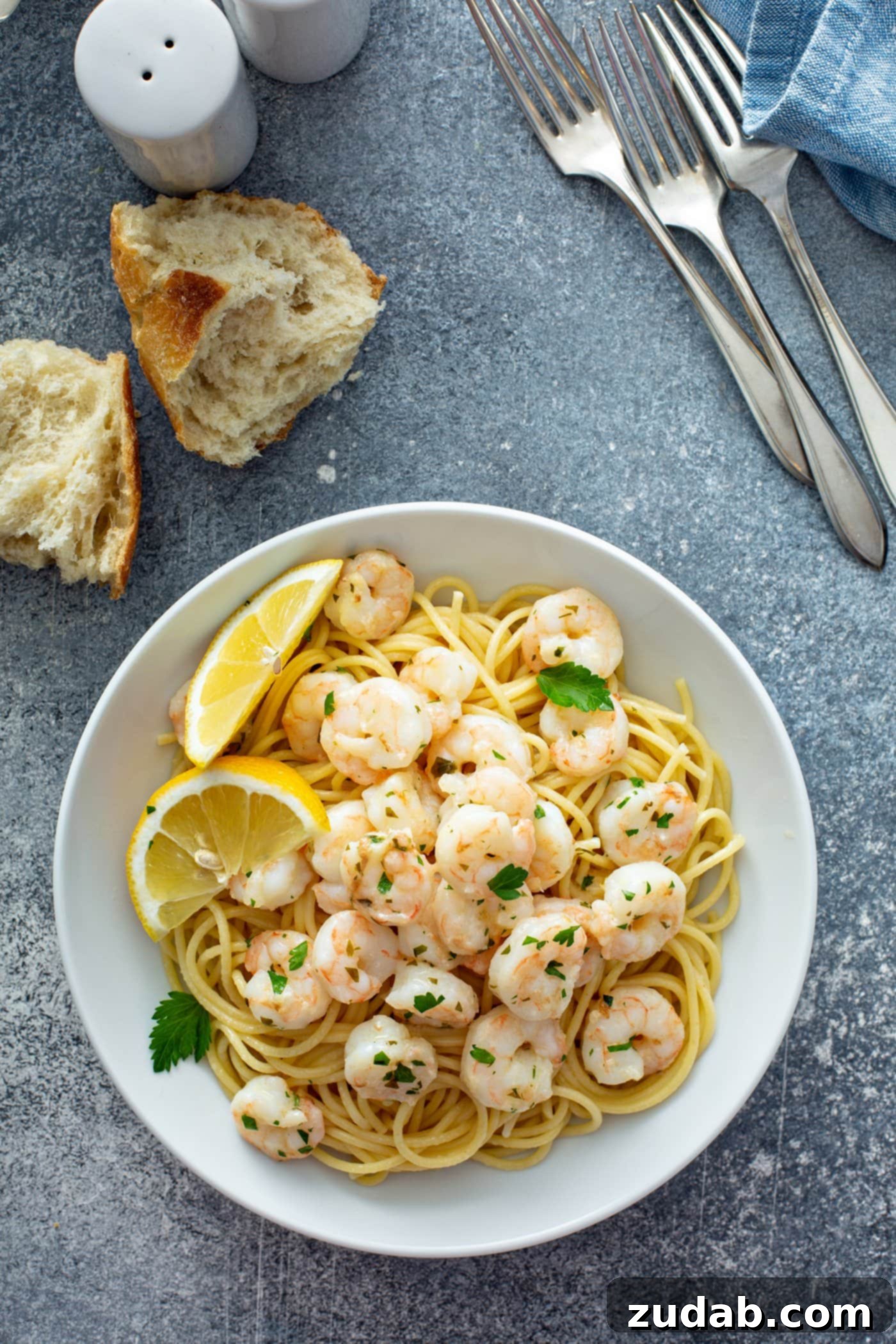 Reheating Shrimp A deep glass bowl filled with succulent lemon garlic shrimp nestled over a bed of pasta. Two bright lemon wedges rest gracefully on the rim of the bowl, and the entire dish is beautifully sprinkled with fresh parsley. In the background, pieces of torn rustic bread and three forks invite diners to partake in this delicious and easy seafood meal.