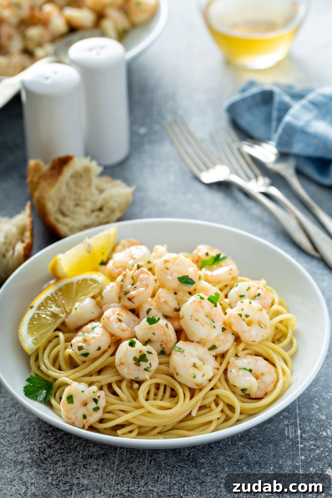 Lemon Garlic Shrimp Tips A delicious serving plate of lemon garlic shrimp elegantly presented over a bed of pasta. Bright lemon wedges garnish the plate, complemented by three forks, salt and pepper shakers, and rustic bread pieces artfully arranged on the table, inviting a delightful dining experience.