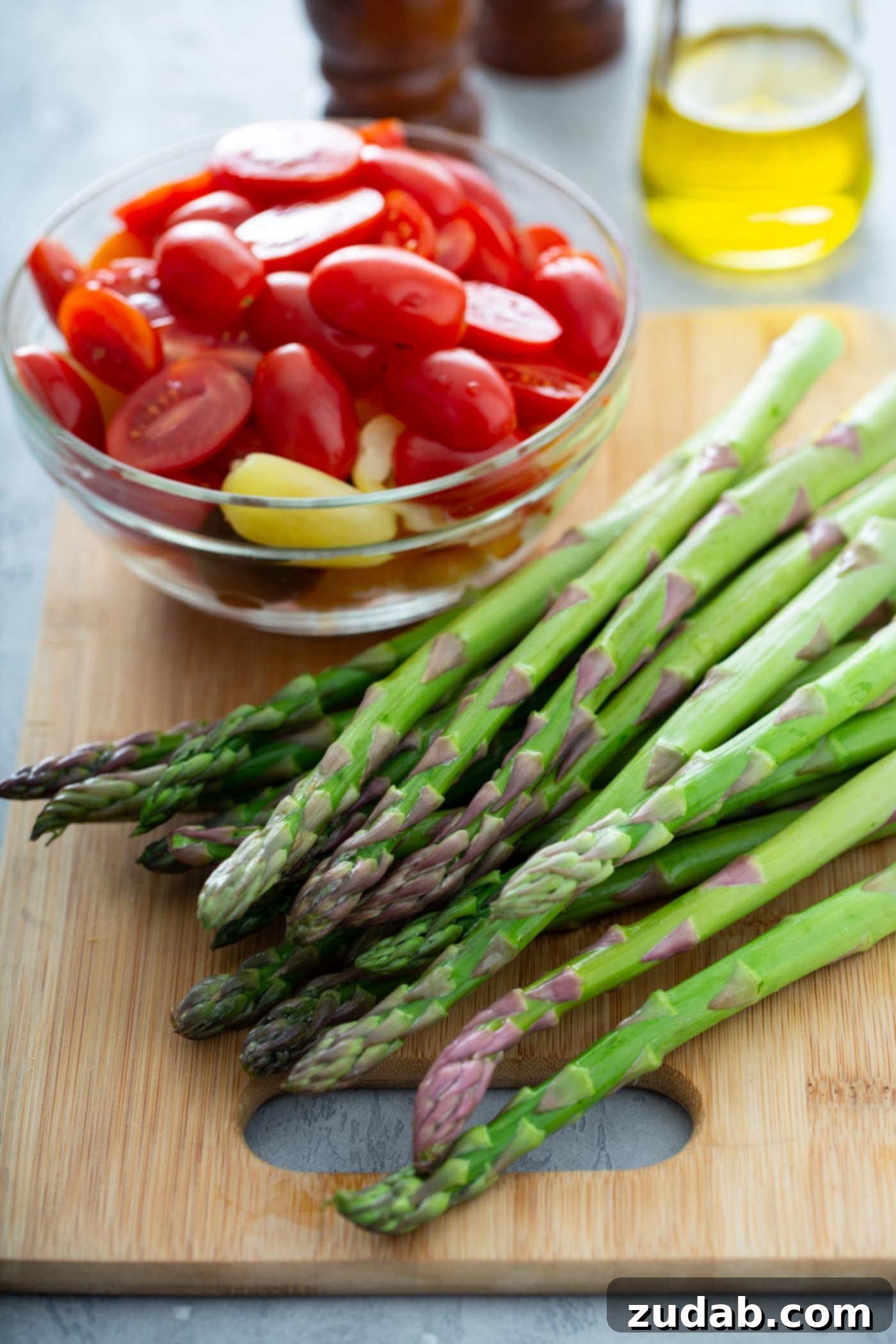 Caramelized Balsamic Asparagus and Tomatoes 3 A bunch of fresh asparagus on a wood cutting board. There is a glass bowl of sliced red and yellow heirloom tomatoes next to the asparagus. A glass container of olive oil is in the background. These fresh ingredients are essential for creating the perfect roasted vegetable dish.