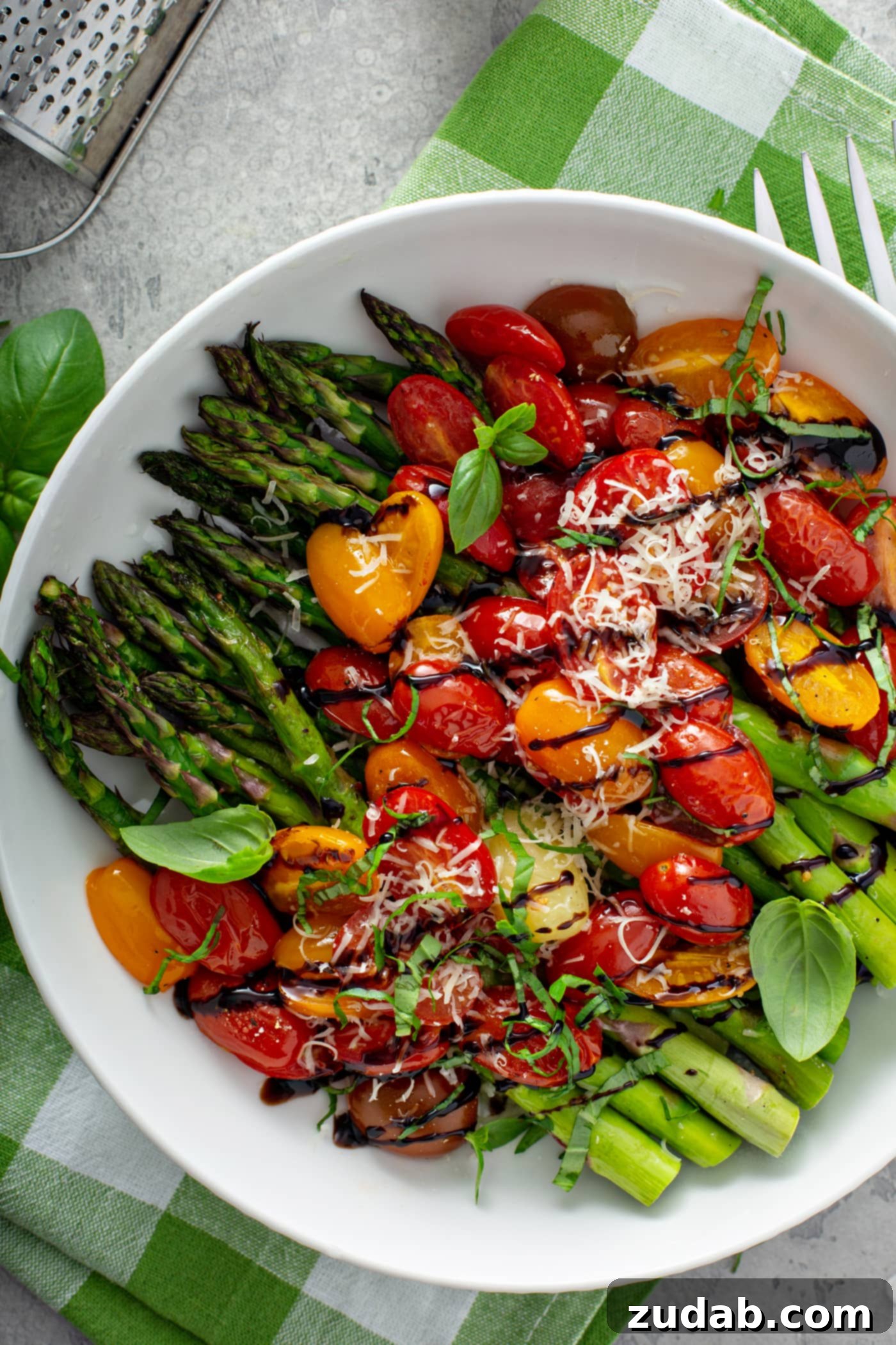 Caramelized Balsamic Asparagus and Tomatoes 5 A serving dish with balsamic glazed roasted tomatoes and asparagus. The vibrant green roasted asparagus is covered with sliced red and yellow tomatoes with a drizzle of balsamic glaze over the top. Grated Parmesan cheese is sprinkled over it all. The serving dish is sitting on a green and white checked cloth, making for a rustic and appealing presentation.