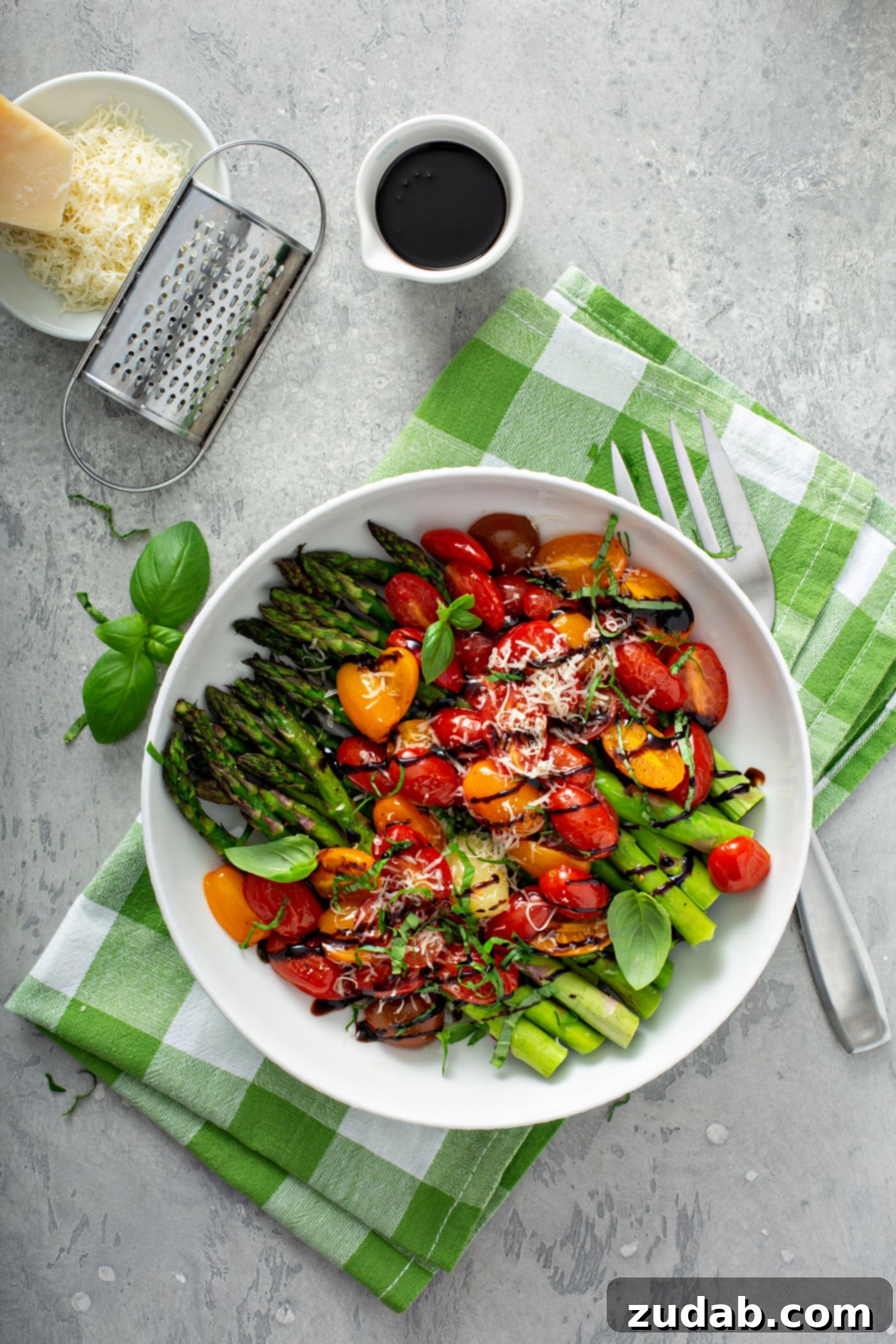 Caramelized Balsamic Asparagus and Tomatoes 7 A serving dish of balsamic glazed tomatoes and asparagus. The bright green asparagus and sliced red and yellow heirloom tomatoes are drizzled with balsamic glaze and sprinkled with grated Parmesan cheese. The serving dish is sitting on a green and white checked cloth. There is a fork beside the dish and a cup of balsamic glaze and a dish of grated cheese and a cheese grater in the background, showcasing the garnishes and components of the dish.