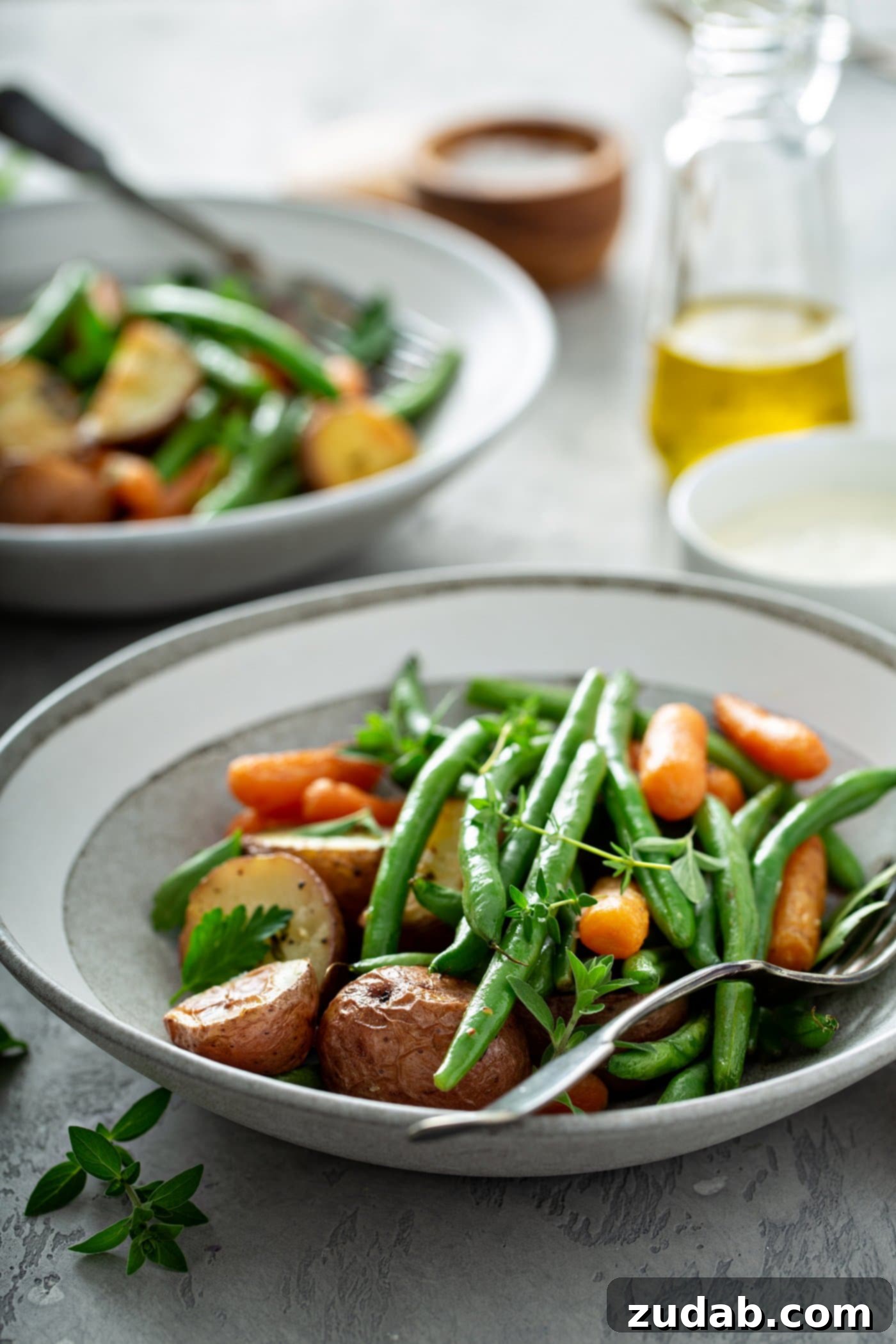 A dinner plate filled with garlic herb roasted potatoes, carrots, and green beans. A fork is resting next to the vegetables. A Serving platter of roasted vegetables, a small bowl of salt, a glass container of oil and
