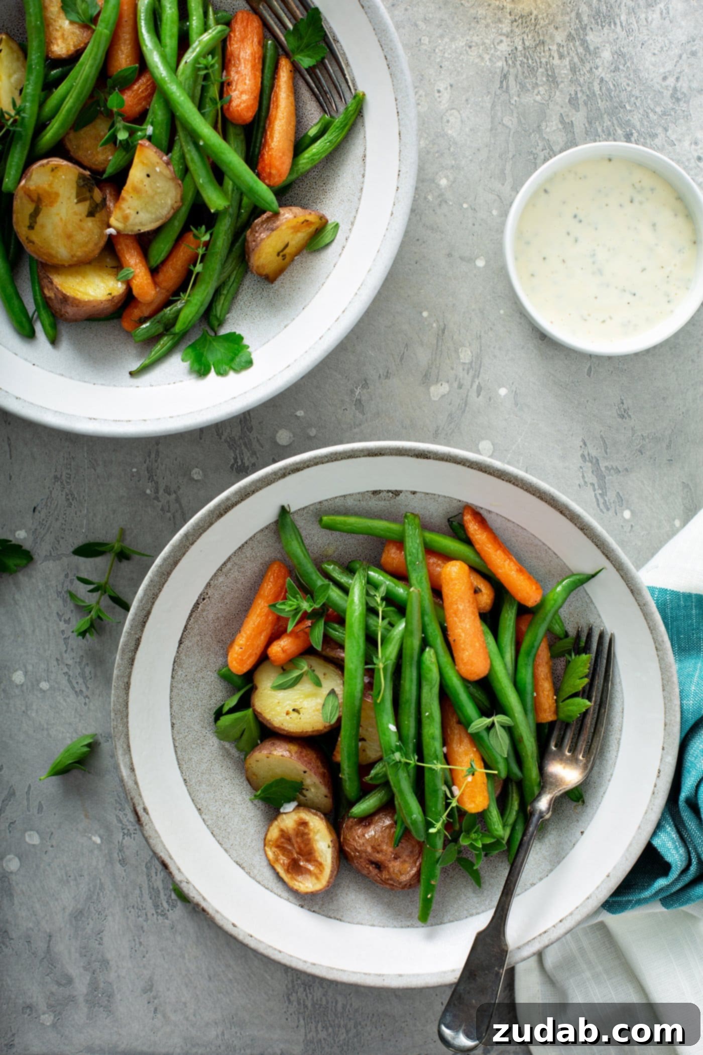 Two dinner plates filled with garlic herb roasted potatoes, carrots and green beans. Fresh parsley and thyme are sprinkled on top and on the table. A napkin is next to one plate, and a small bowl of salt is in the right corner.