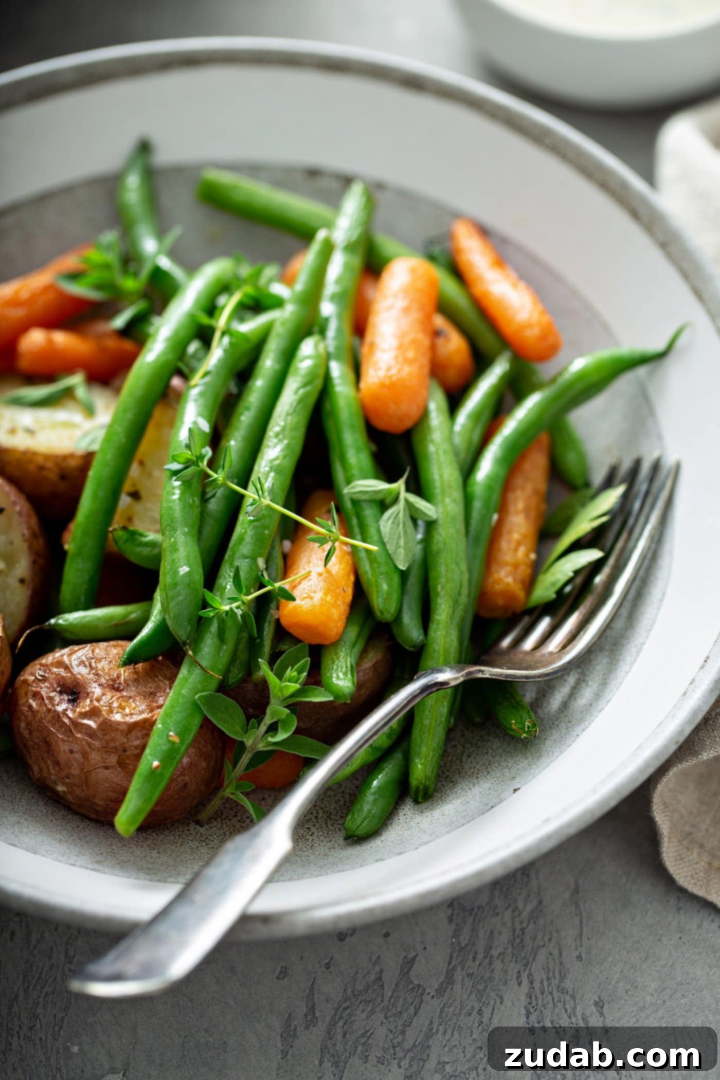 A dinner plate of garlic herb roasted potatoes, carrots and green beans. A fork is laying on the plate next to the vegetables, and they are topped with fresh thyme.