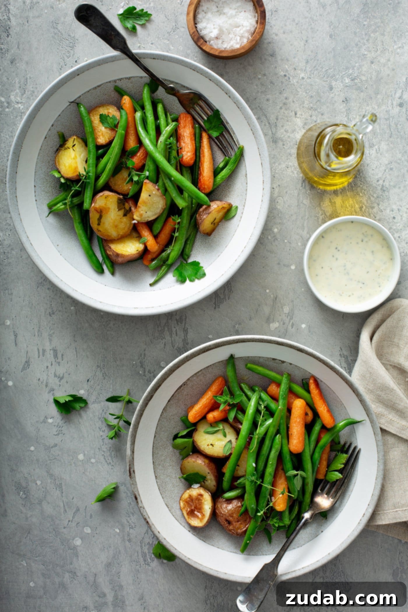 Two dinner plates filled with garlic herb roasted potatoes, carrots and green beans. Fresh thyme and parsley are sprinkled on top. A small container of sauce, a glass container of oil and a napkin are on the table next to the vegetables.