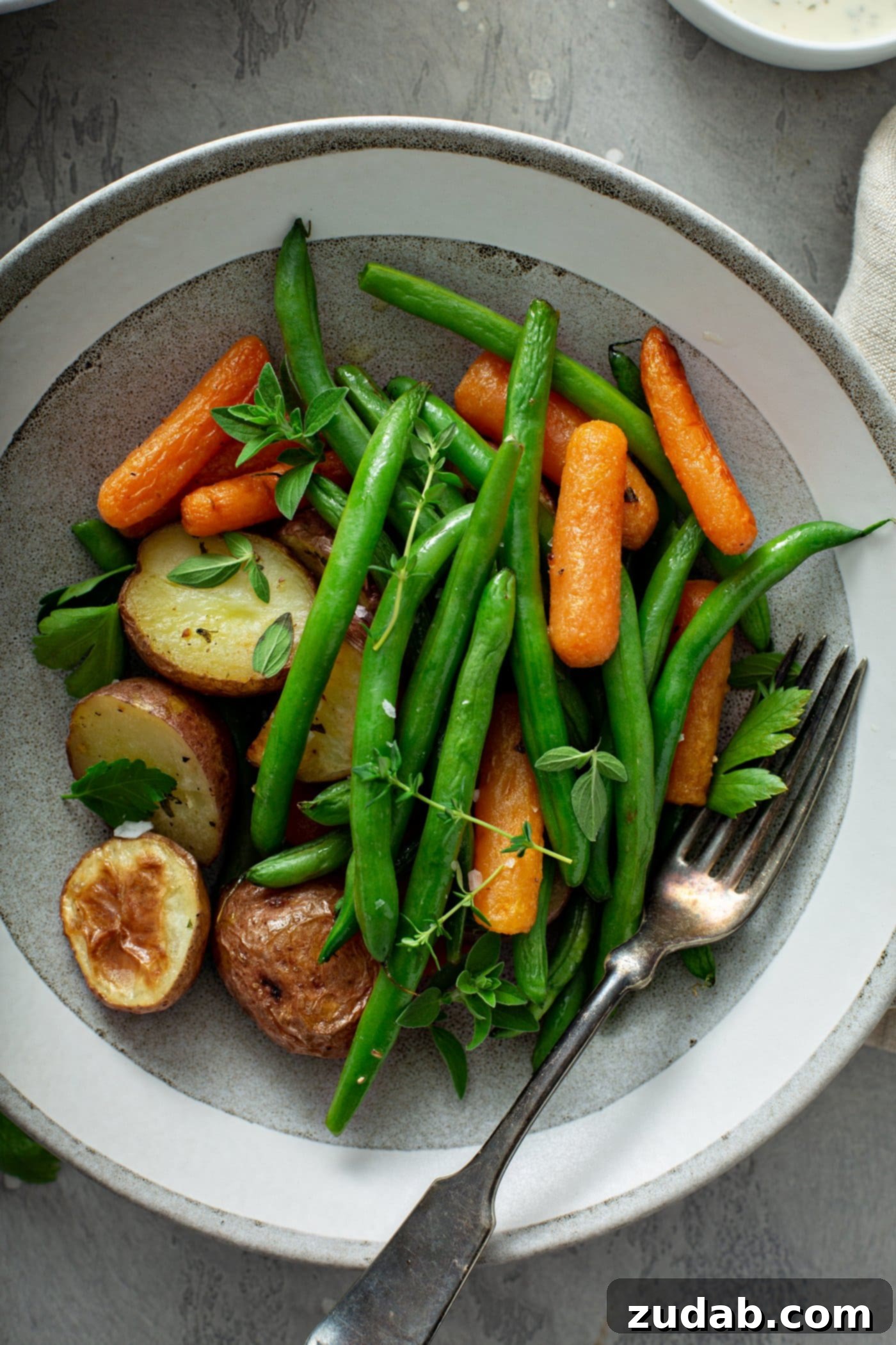 A dinner plate with roasted potatoes, carrots, and green beans. The vegetables are sprinkled with fresh herbs and a fork is resting on the plate next to the vegetables.