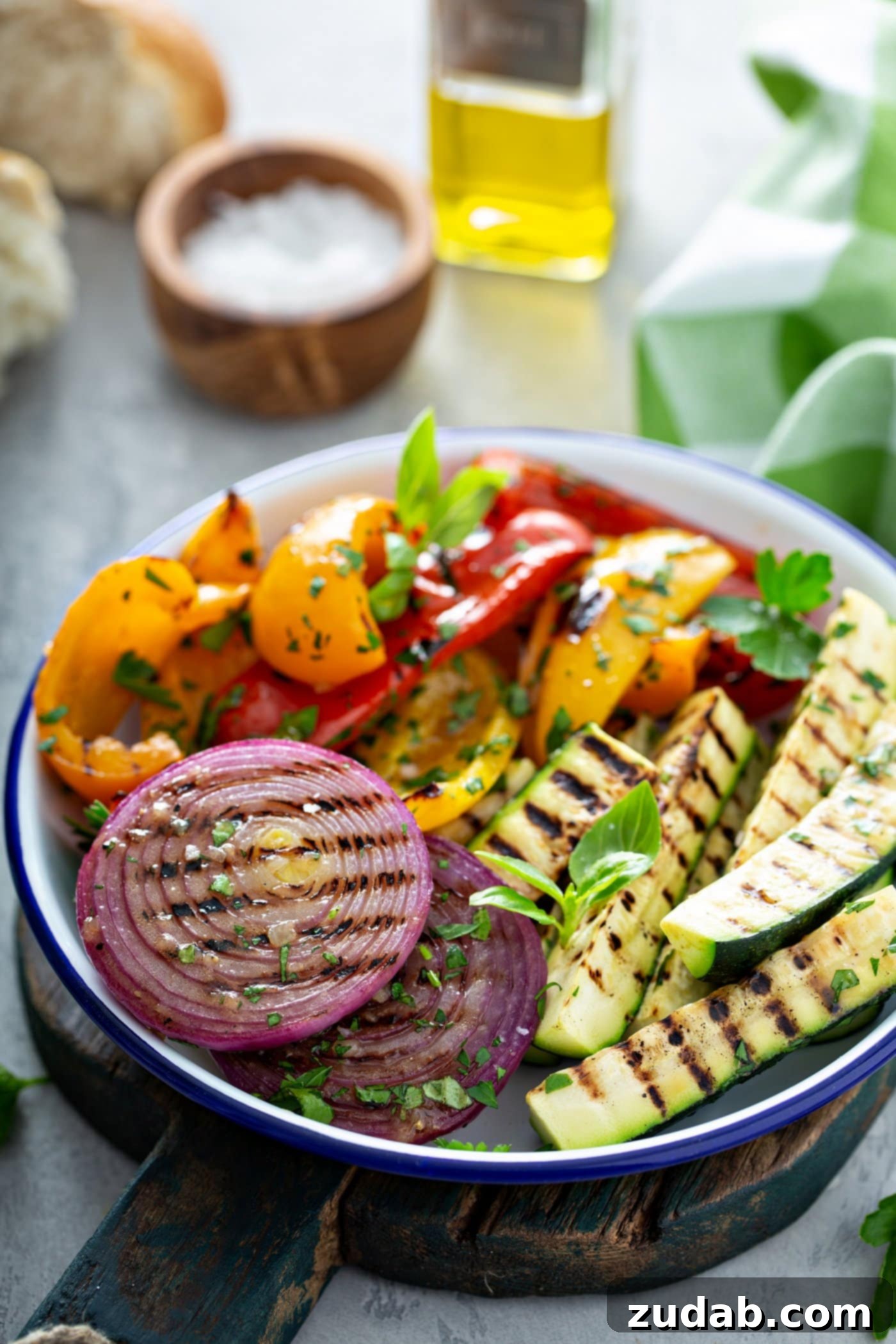Grilled Vegetables with Balsamic Vinaigrette 3 A beautifully arranged serving dish brimming with grilled vegetables including sliced red onions, vibrant red and yellow bell peppers, and tender zucchini, all displaying tempting grill marks and garnished with fresh parsley. In the background, there's a rustic wooden salt cellar, a glass oil dispenser, and a charming green and white checked cloth, setting a perfect scene for a summer meal.
