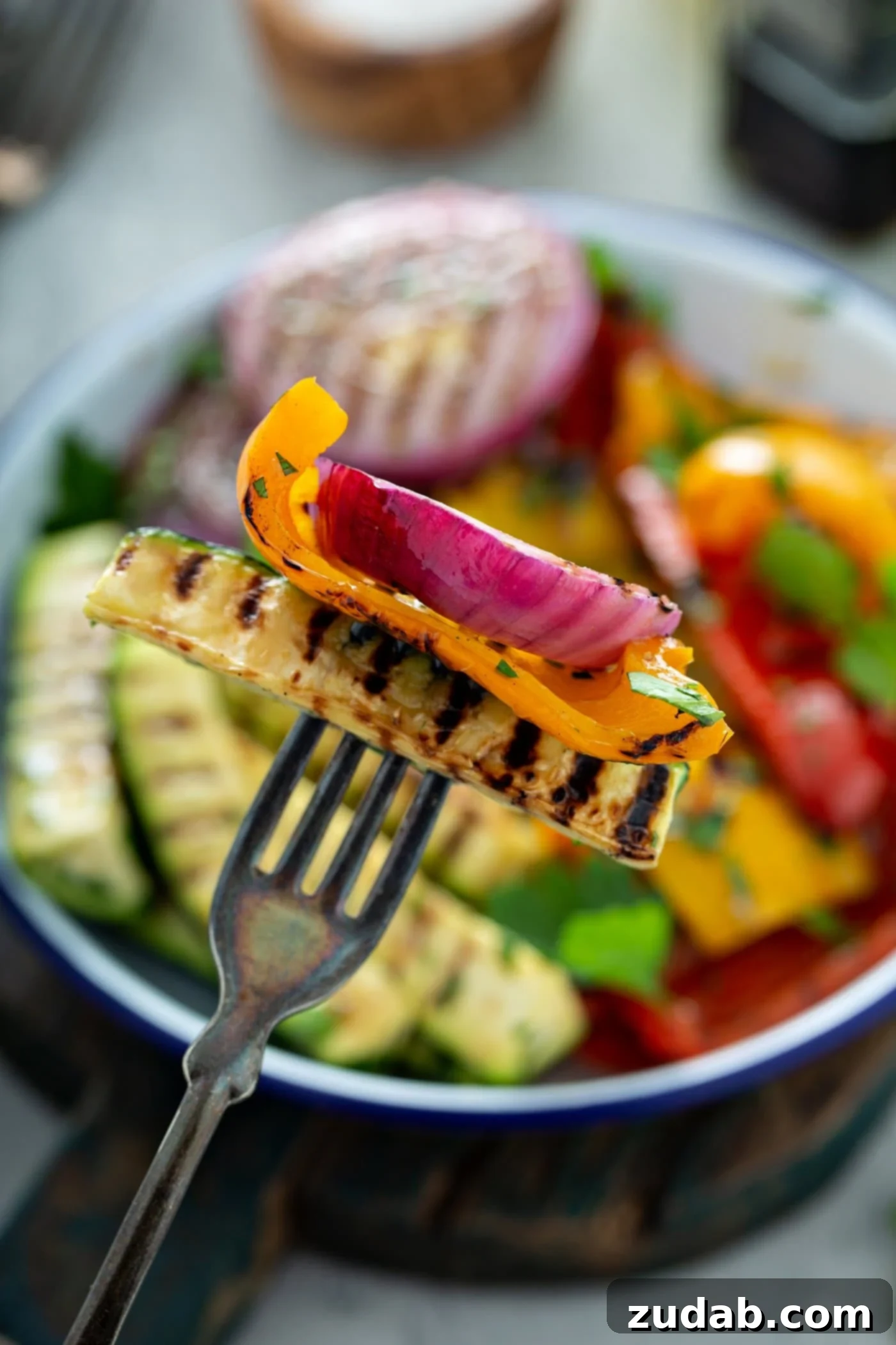 Grilled Vegetables with Balsamic Vinaigrette 4 A fork delicately pierces a piece of grilled zucchini, yellow bell pepper, and red onion, showcasing the beautiful grill marks. In the soft-focus background, a serving dish of more grilled vegetables hints at the feast to come.
