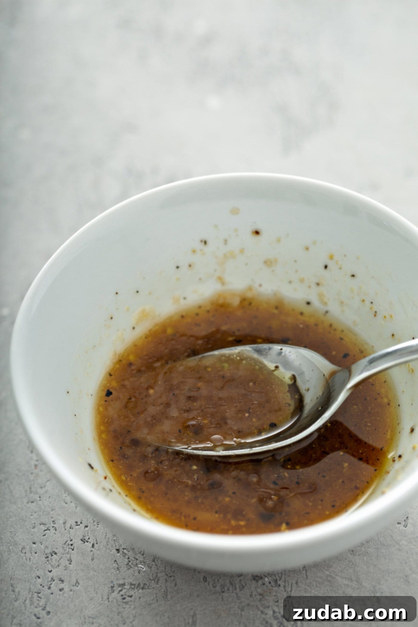 Grilled Vegetables with Balsamic Vinaigrette 8 A pristine white bowl holds a generous portion of glossy balsamic glaze, with a spoon resting inside, ready to be drizzled over dishes.