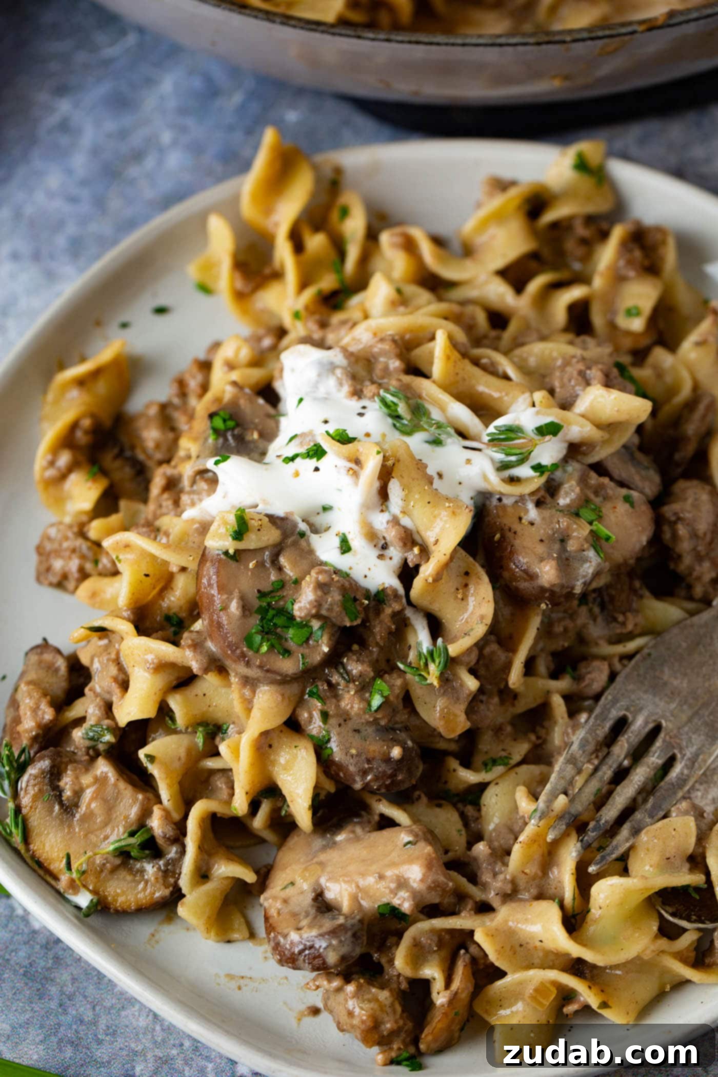 Ground Beef Stroganoff Ground beef stroganoff on a dinner plate. A fork rests on the plate next to the noodles and ground beef stroganoff with a spoonful of sour cream on top.