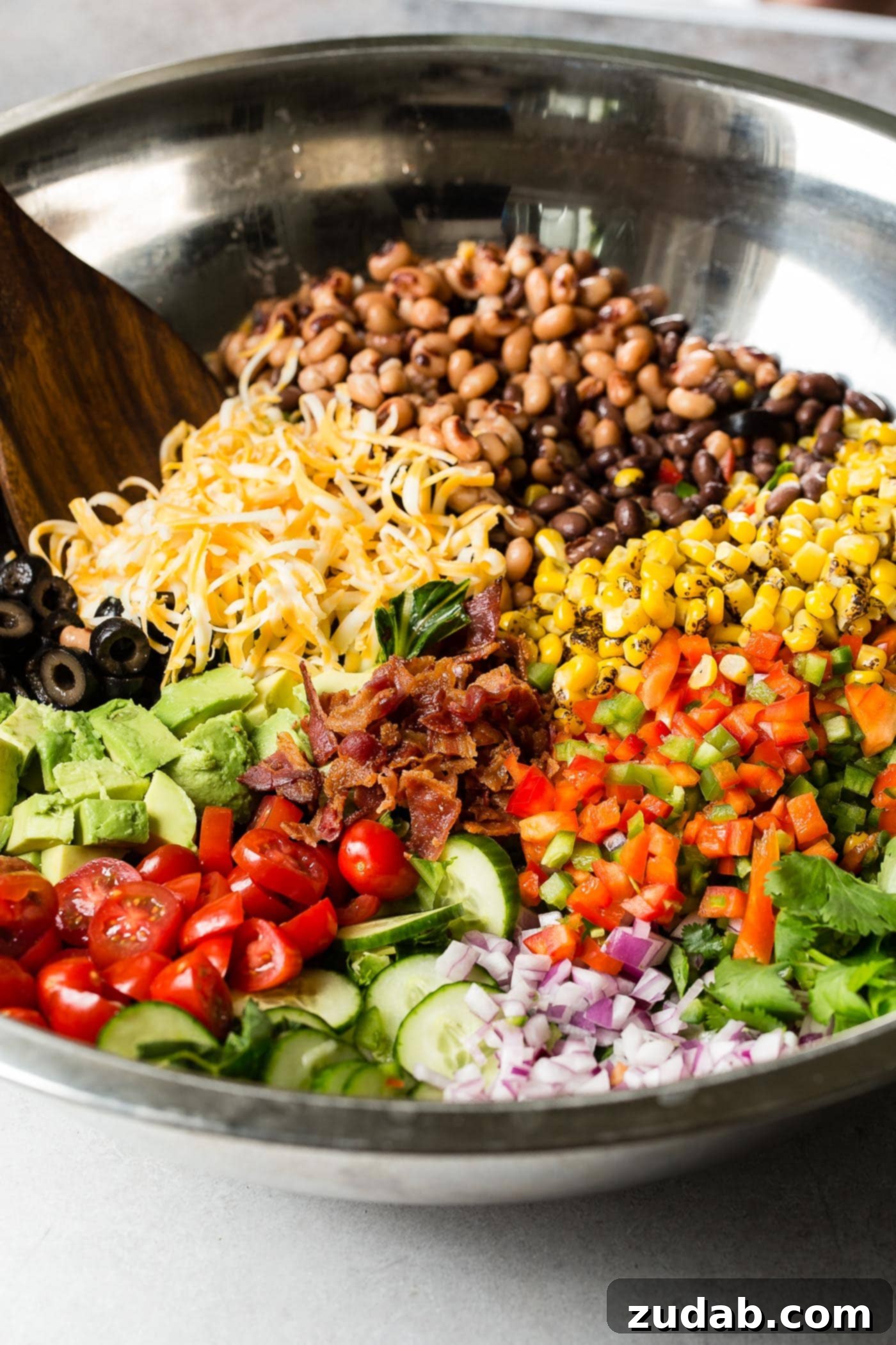 Chuckwagon Salad 4 Sections of chopped tomatoes, avocados, red onion, corn, cilantro, beans, shredded cheese, olives, peppers, bacon, and peas in a glass bowl with a wooden spoon.