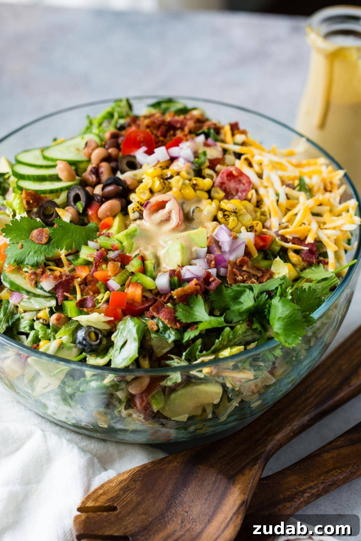 Chuckwagon Salad 6 Cowboy salad in a glass serving bowl all ready to be served. It has sliced cucumbers, chopped lettuce, avocados, peppers, bacon, and olives and shredded cheese. It is topped with dressing.