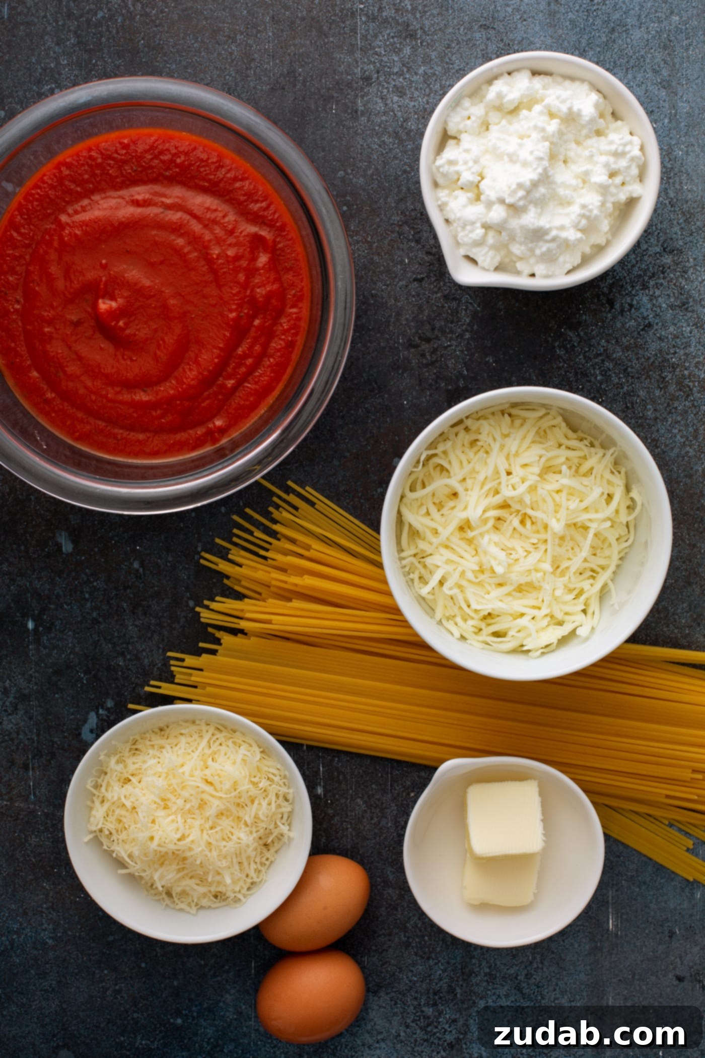 Baked Spaghetti Pie A small, rustic wooden bowl containing all the essential ingredients for preparing baked spaghetti pie. Inside, there's a neat handful of uncooked spaghetti pasta, a vibrant red bowl overflowing with rich spaghetti sauce, and three small, clear containers holding an assortment of cheeses: shredded mozzarella cheese, creamy cottage cheese, and finely grated Parmesan cheese. This image perfectly illustrates the simple yet delicious components of the dish.