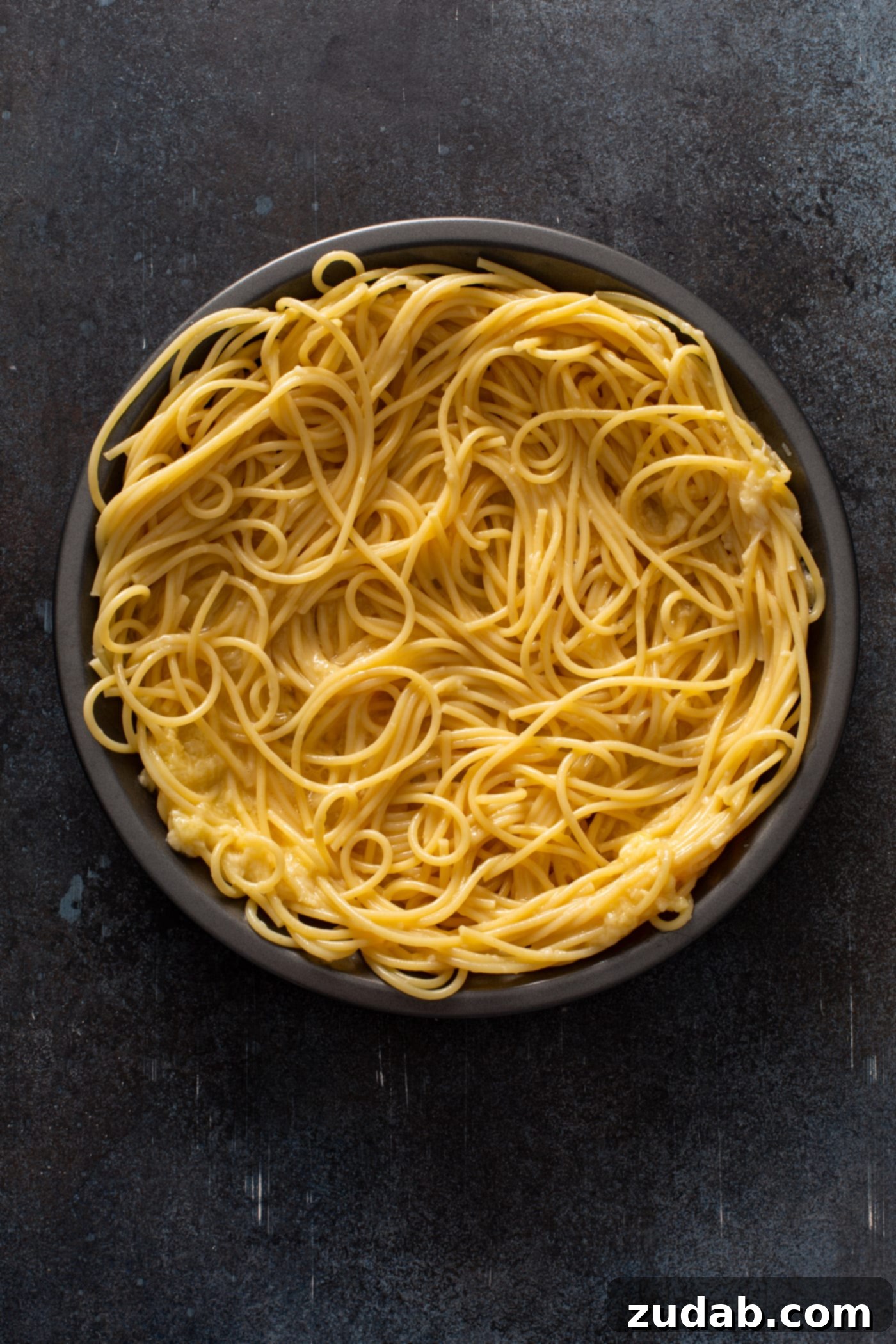 Baked Spaghetti Pie A deep pie dish containing a generous portion of cooked spaghetti noodles, ready for the next delicious layers. The noodles are evenly spread, forming the base of what will become a comforting baked spaghetti pie. The image focuses on the pasta, highlighting its texture and preparation stage.