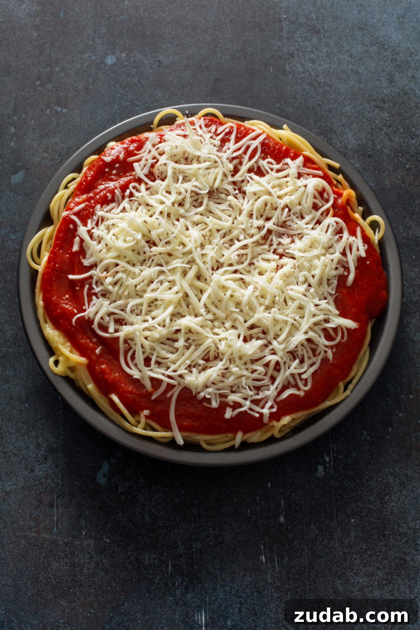Baked Spaghetti Pie A deep pie dish filled with cooked spaghetti, generously topped with a vibrant spaghetti sauce and a blanket of shredded cheeses, ready to be baked. The rich red sauce contrasts beautifully with the white cheese, hinting at the delicious flavors within this comforting Italian-American dish.