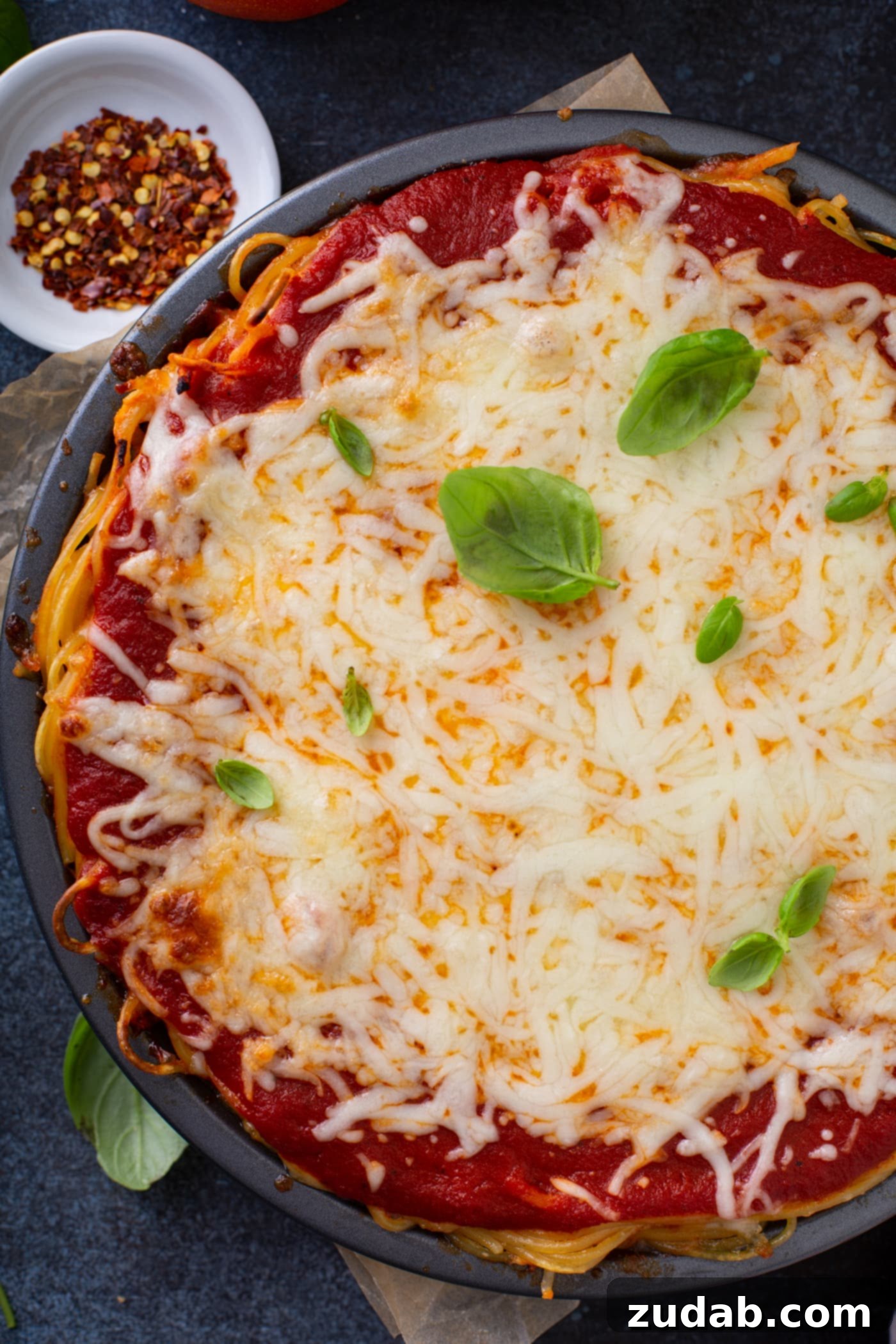 Baked Spaghetti Pie A vibrant top-down view of a baked spaghetti pie, generously filled with golden-brown spaghetti pasta, rich spaghetti sauce, and a topping of perfectly melted cheeses. A few fresh basil leaves are scattered over the surface, adding a pop of color and freshness. In the blurred background, a small, clear bowl of dried red pepper flakes sits, hinting at an option to add a touch of spice to this comforting dish.