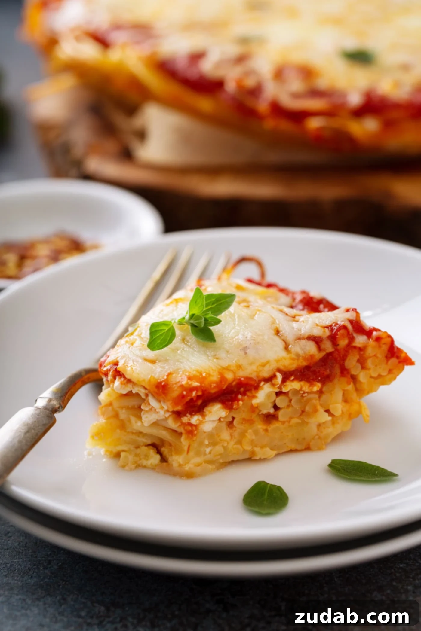 Baked Spaghetti Pie A perfect slice of baked spaghetti pie meticulously placed on a white dinner plate. The slice clearly displays distinct layers of seasoned spaghetti, rich spaghetti sauce, and beautifully melted shredded cheese. A fork rests invitingly next to the spaghetti pie, and a few fresh basil leaves are artfully scattered both on the pie and the plate, adding a touch of color and freshness. In the background, subtly blurred, a small container of red pepper flakes and the remaining baked spaghetti pie in its dish are visible, creating a complete and tempting culinary scene.