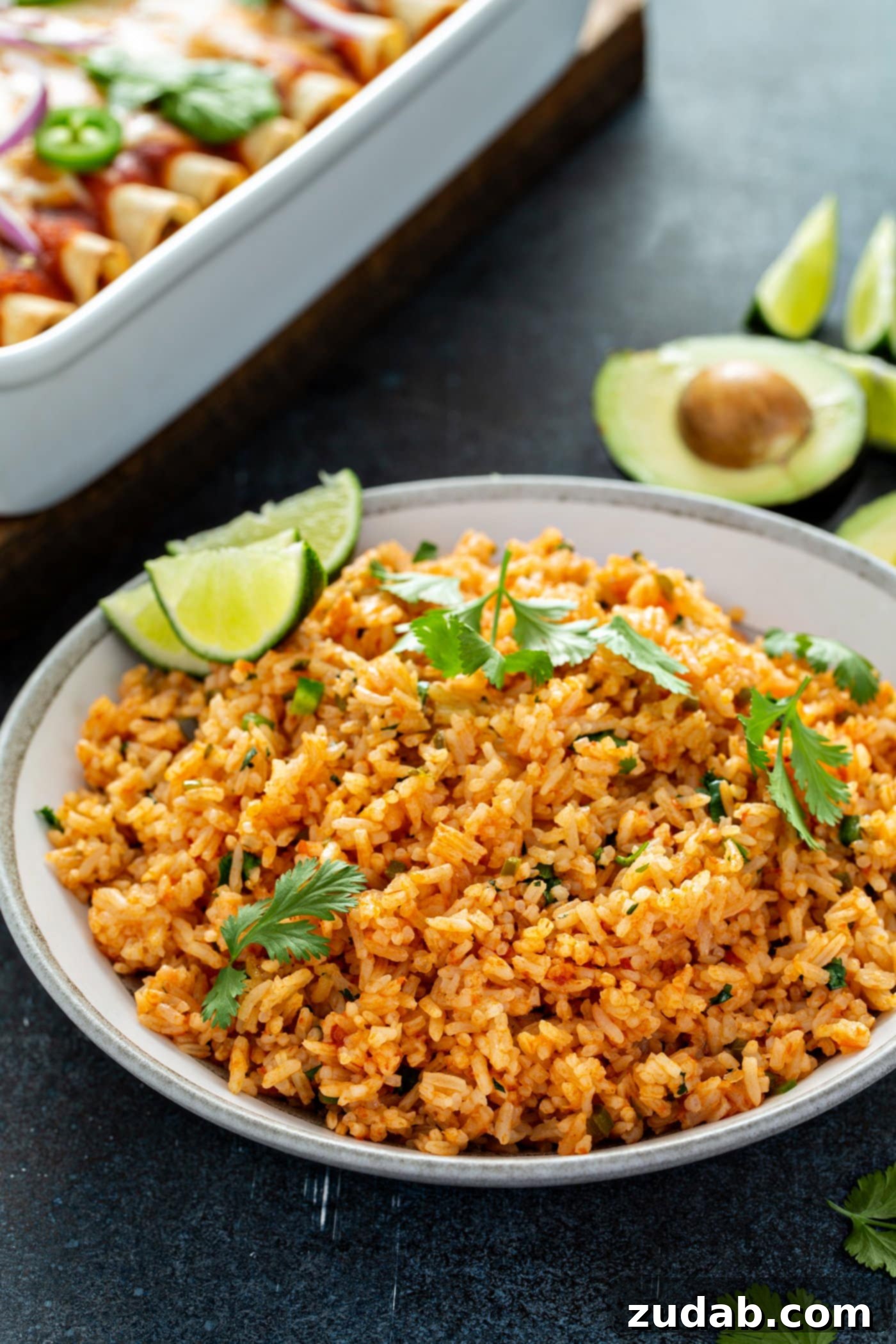 A bowl of vibrant Spanish rice, garnished with fresh cilantro and lime wedges, with enchiladas and avocado in the background.