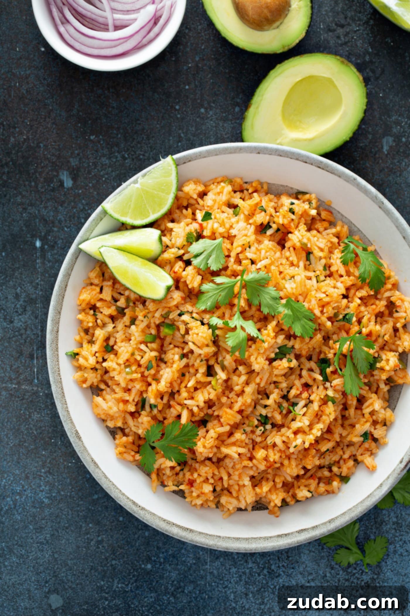 A serving bowl of Spanish rice topped with cilantro leaves and three lime wedges. There is a halved avocado in the background.