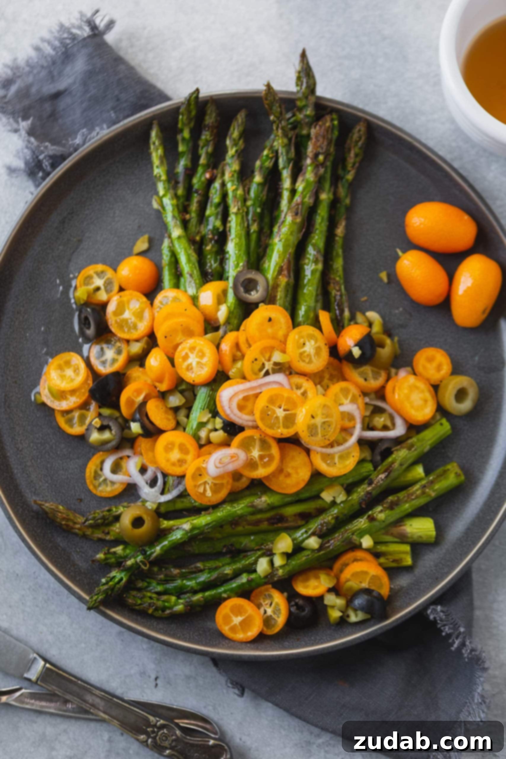 A vibrant photo of perfectly roasted asparagus spears on a charcoal gray serving dish, beautifully adorned with bright orange sliced kumquats, delicate sliced shallots, and a scattering of both black and green olives. The dish promises a symphony of flavors and textures.