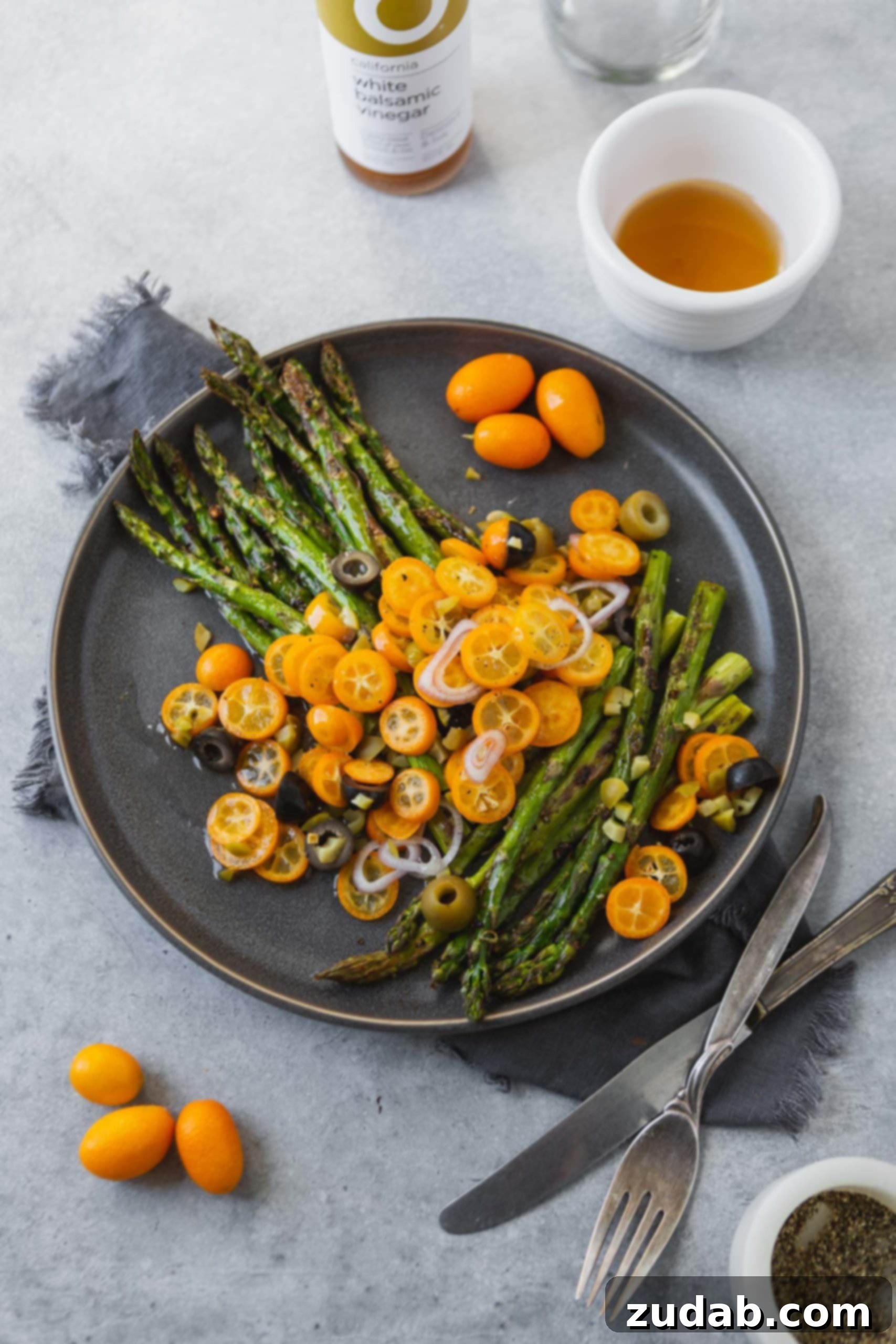 Another close-up shot of the roasted asparagus with bright orange kumquat slices, shallots, and a mix of black and green olives, highlighting the fresh, appealing presentation of the dish.
