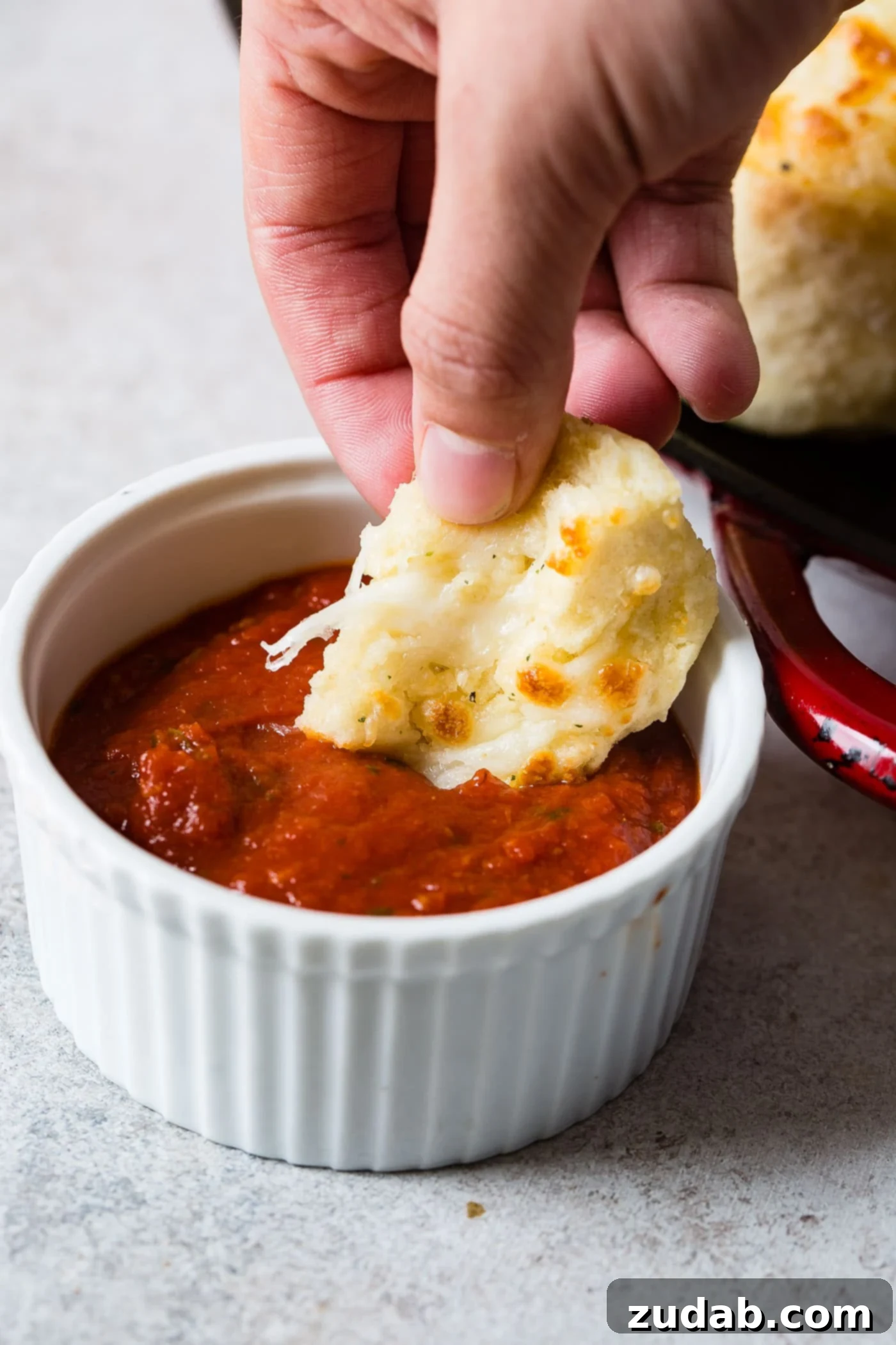 Golden Cheesy Perfection 4 A piece of cheese doodle cheesy bread being dipped in a a small white bowl of marinara sauce. A tray of cheese doodles sits next to the sauce.