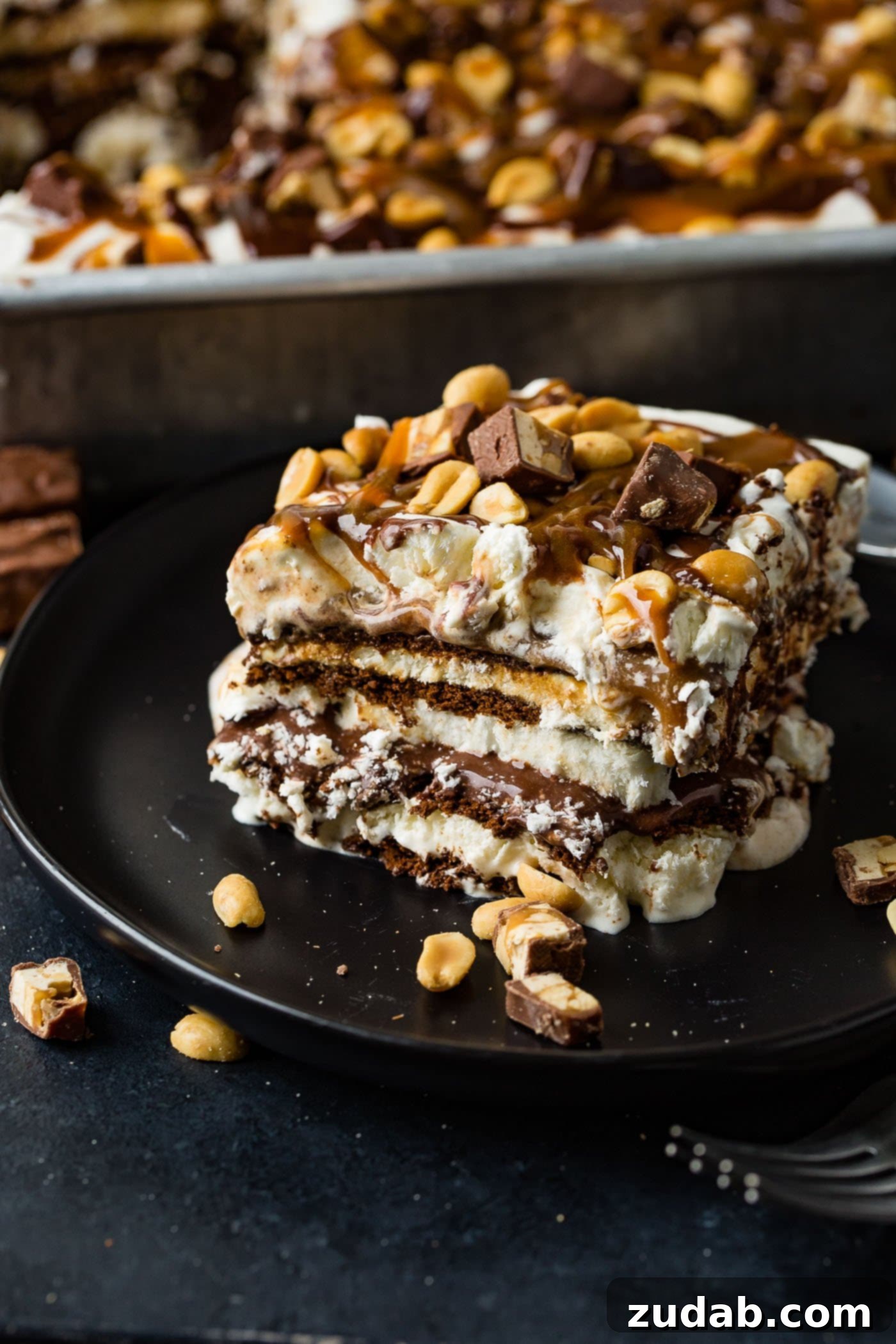 Ultimate Snickers Ice Cream Cake 2 A square piece of snickers ice cream cake on a brown dessert plate. There are layers of ice cream, chocolate cake, fudge, caramel and peanuts. There is fudge and whipped cream on top and salted peanuts are sprinkled over the whole thing. A pan of snickers ice cream cake is in the background.
