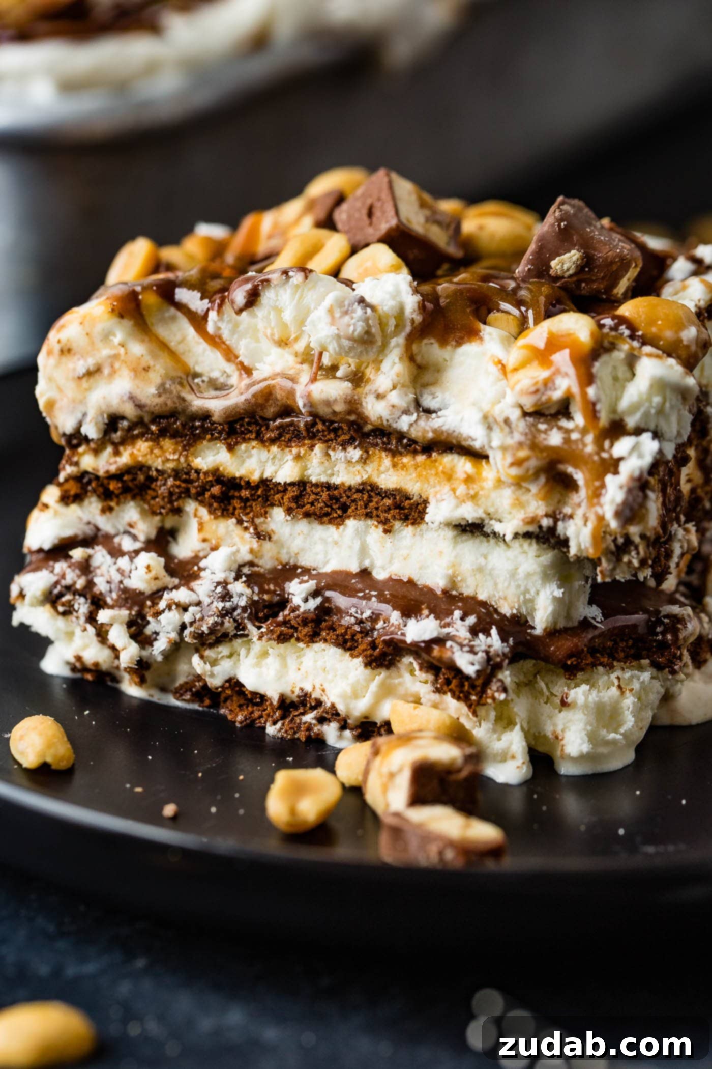 Ultimate Snickers Ice Cream Cake 3 A square piece of snickers ice cream cake. There are layers of cake, ice cream, hot fudge, caramel sauce, snickers candy bars and whipped cream. The cake is topped with whipped cream, drizzles of hot fudge and caramel sauce and salted peanuts.