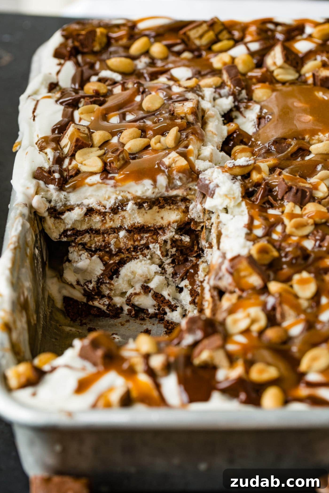 Ultimate Snickers Ice Cream Cake 4 A pan of snickers ice cream cake with a piece removed. There are layers of ice cream, cake, fudge, caramel, whipped cream and candy bars. The top is drizzled with hot fudge, caramel topping and salted peanuts.