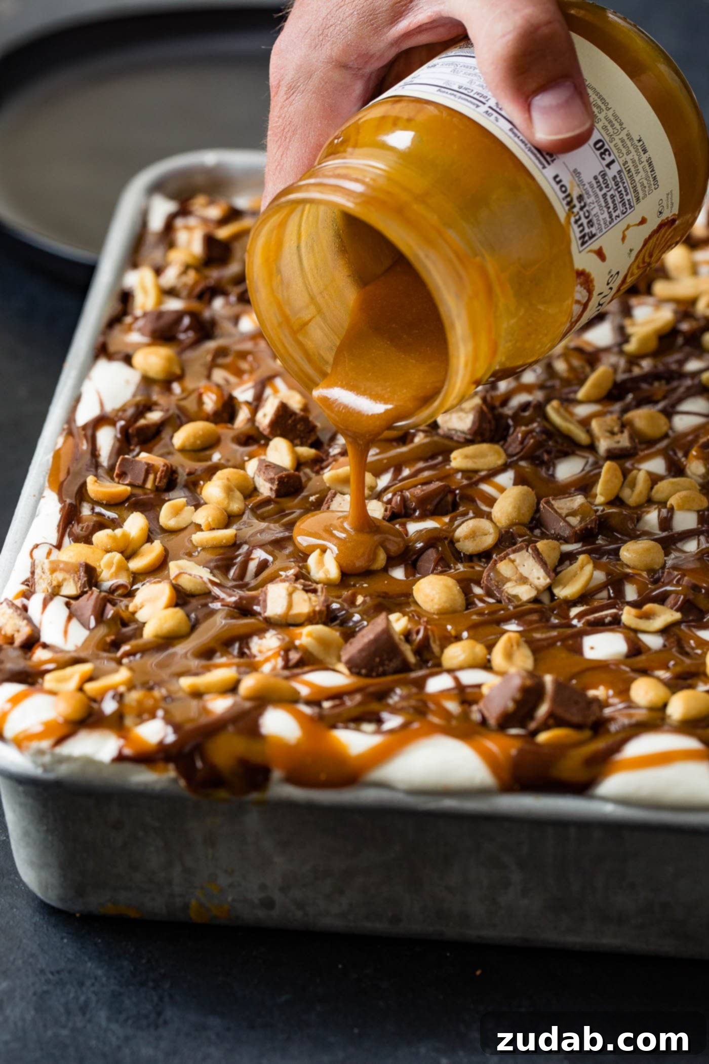 Ultimate Snickers Ice Cream Cake 5 A jar of melted caramel topping being poured over the top of a snickers ice cream cake. The top of the cake is covered with whipped cream, drizzles of hot fudge, and salted peanuts.