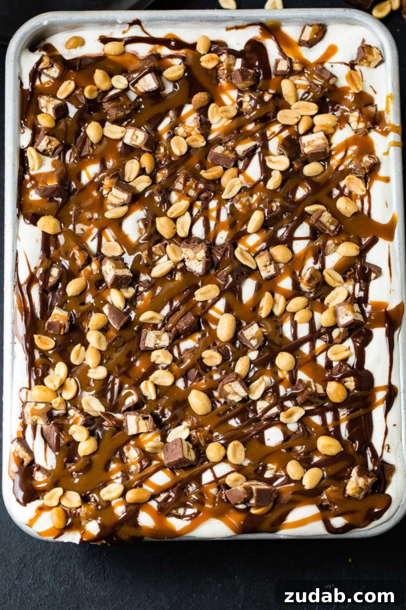 Ultimate Snickers Ice Cream Cake 6 A pan of snickers ice cream cake. The top is covered with whipped cream, hot fudge, drizzles of caramel sauce, chopped snickers bars and salted peanuts