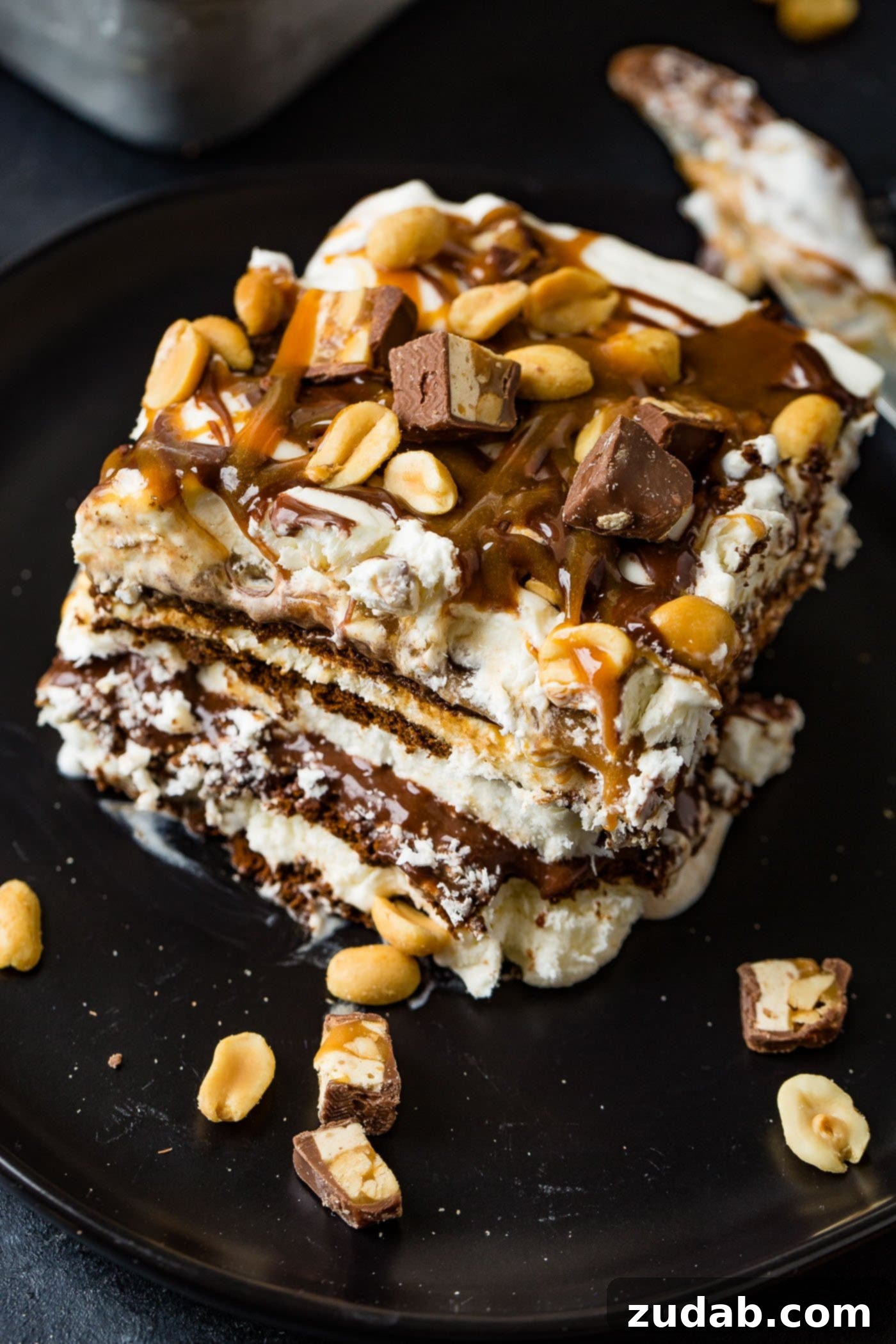 Ultimate Snickers Ice Cream Cake 7 A piece of snickers ice cream cake on a dessert plate. The cake has layers of chocolate cake, ice cream, whipped cream, hot fudge, and caramel sauce. The top is covered with whipped cream, drizzles of hot fudge and caramel sauce, and salted peanuts. Chunks of snickers bars and peanuts are scattered around it.