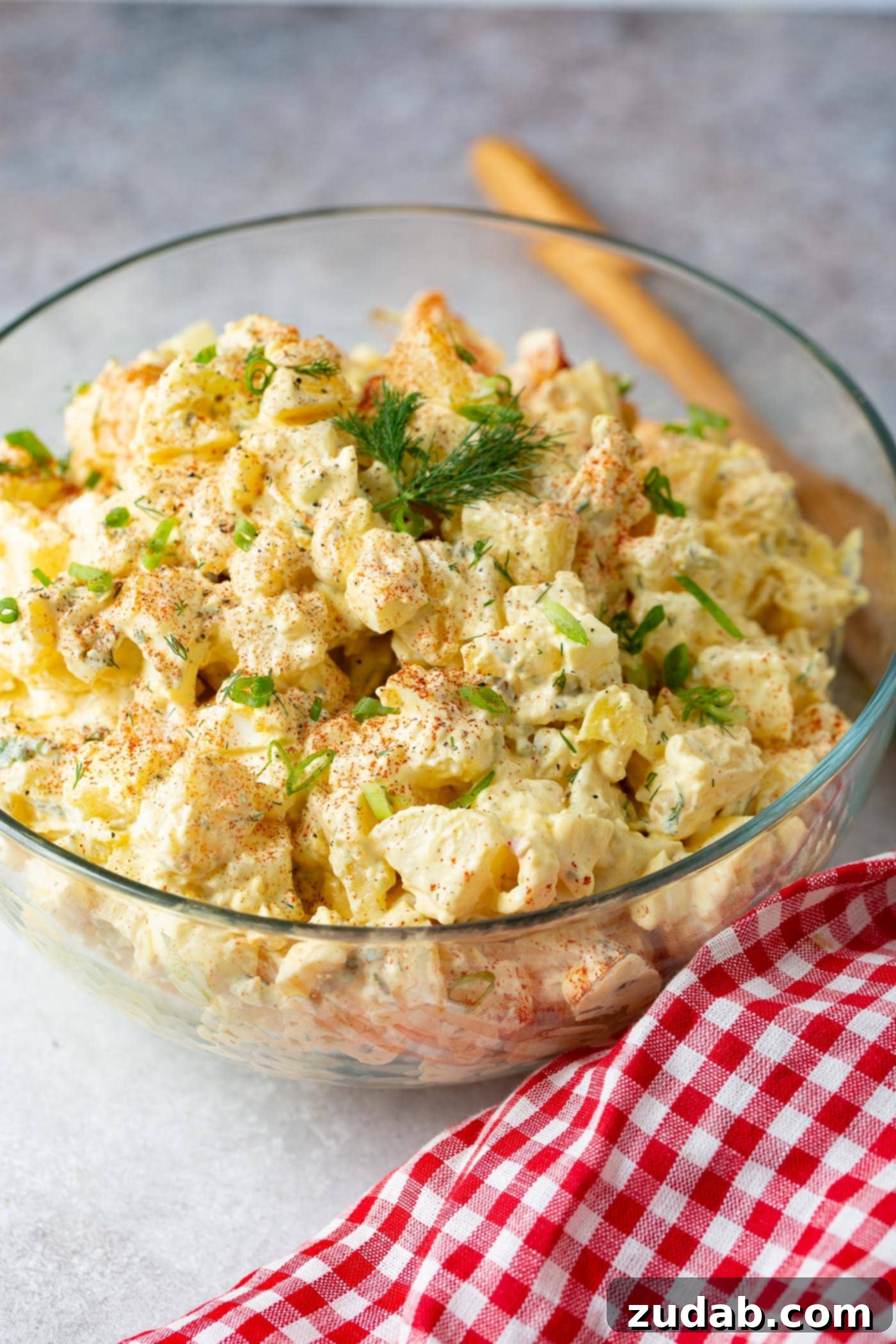 Secrets to the Best Potato Salad Ever 2 A glass serving bowl filled with a BBQ side! There are chunks of chopped eggs, fresh dill, and it is sprinkled with paprika and fresh parsley leaves. The bowl is sitting on a table next to a red checked napkin. A wooden spoon is also laying on the table.