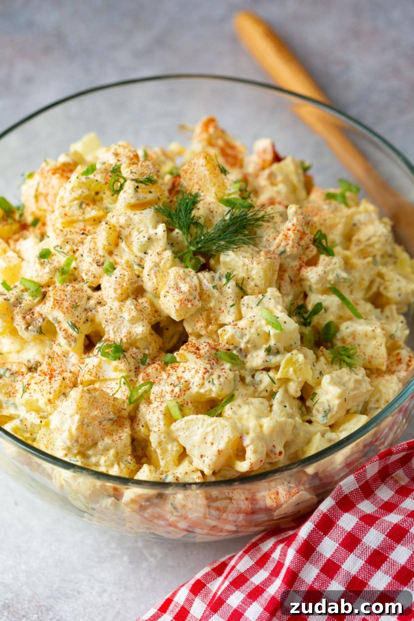 Secrets to the Best Potato Salad Ever 3 A glass serving bowl with this classic BBQ side. It is topped with sprinkled paprika and fresh parsley. The bowl is sitting next to a red checked napkin and a wooden spoon.