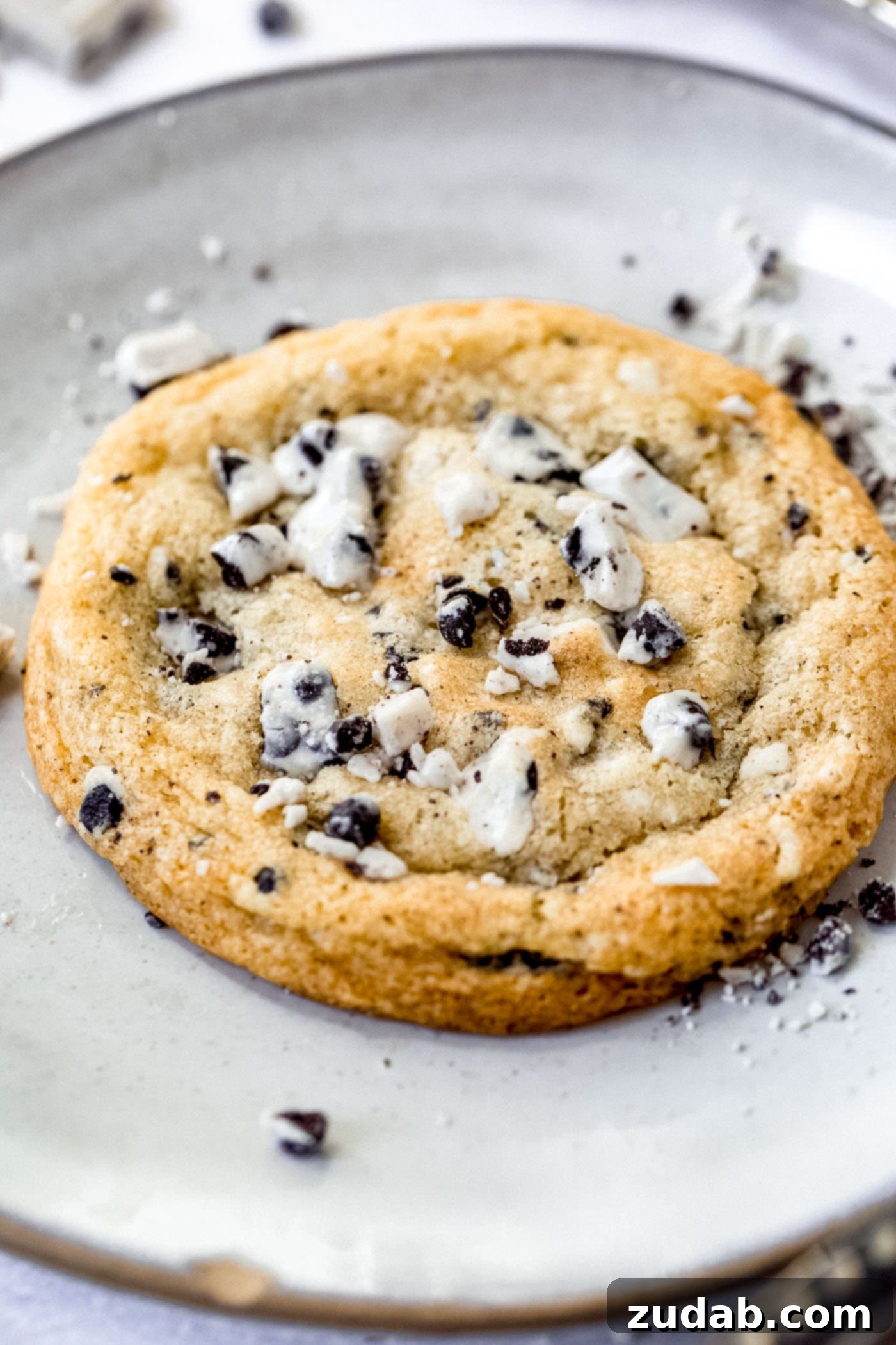 Irresistible Cookies and Cream Cookies A large cookies and cream cookie on a dessert plate. The cookie is generously studded with bits of chocolate cookie and white cream, and there are more cookie pieces scattered around it, highlighting its rich texture.