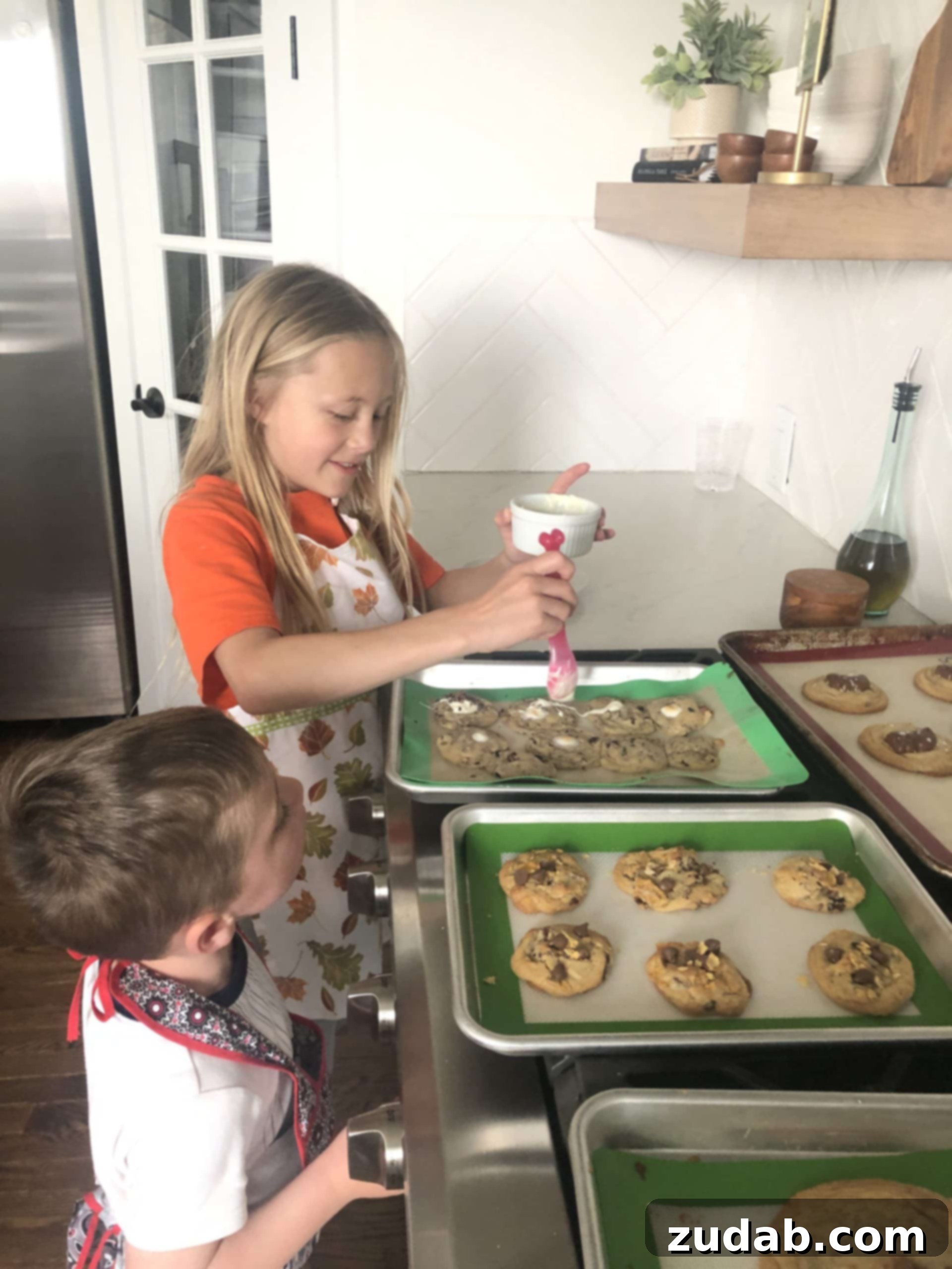Family Baking Fun A photo of a young girl spooning melted white chocolate over chocolate chip cookies with her little brother watching, illustrating a fun family baking moment.