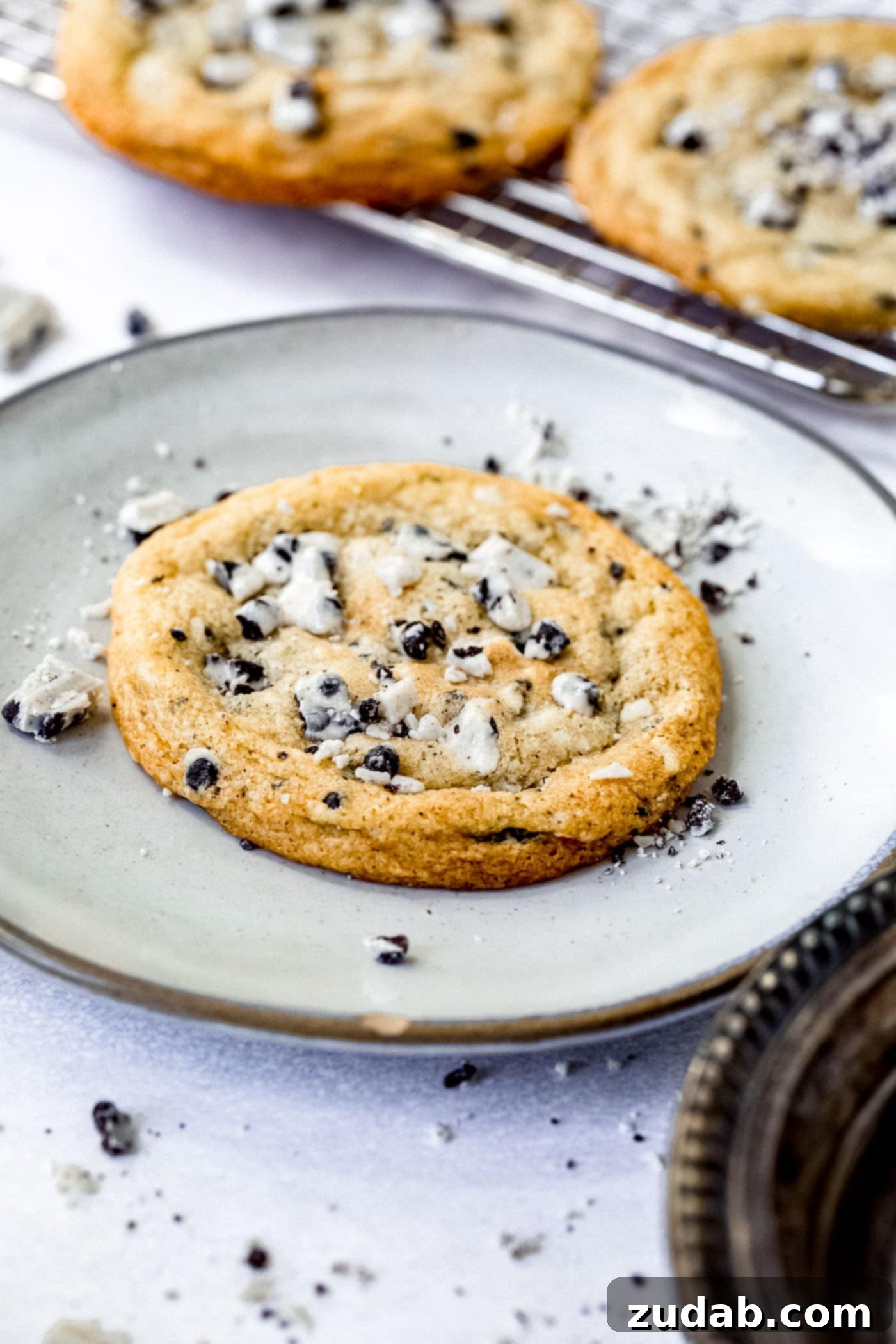 Freshly Baked Cookies and Cream a large cookies and cream cookie on a white dessert plate. The cookie is loaded with bits of cookies and cream candy bar. there are bits of candy bar on the plate and a cooling rack of cookies in the background.