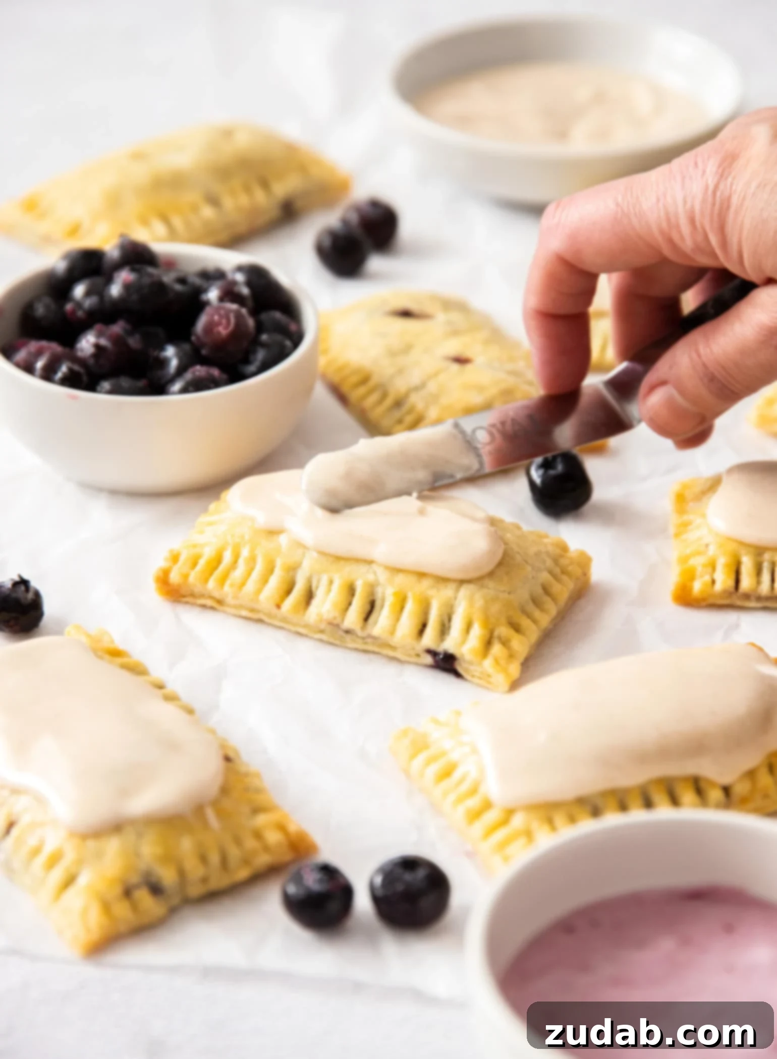 DIY Blueberry Toaster Pastries 6 homemade pop tarts being spread with icing. A small white bowl filled with blueberries to the side.