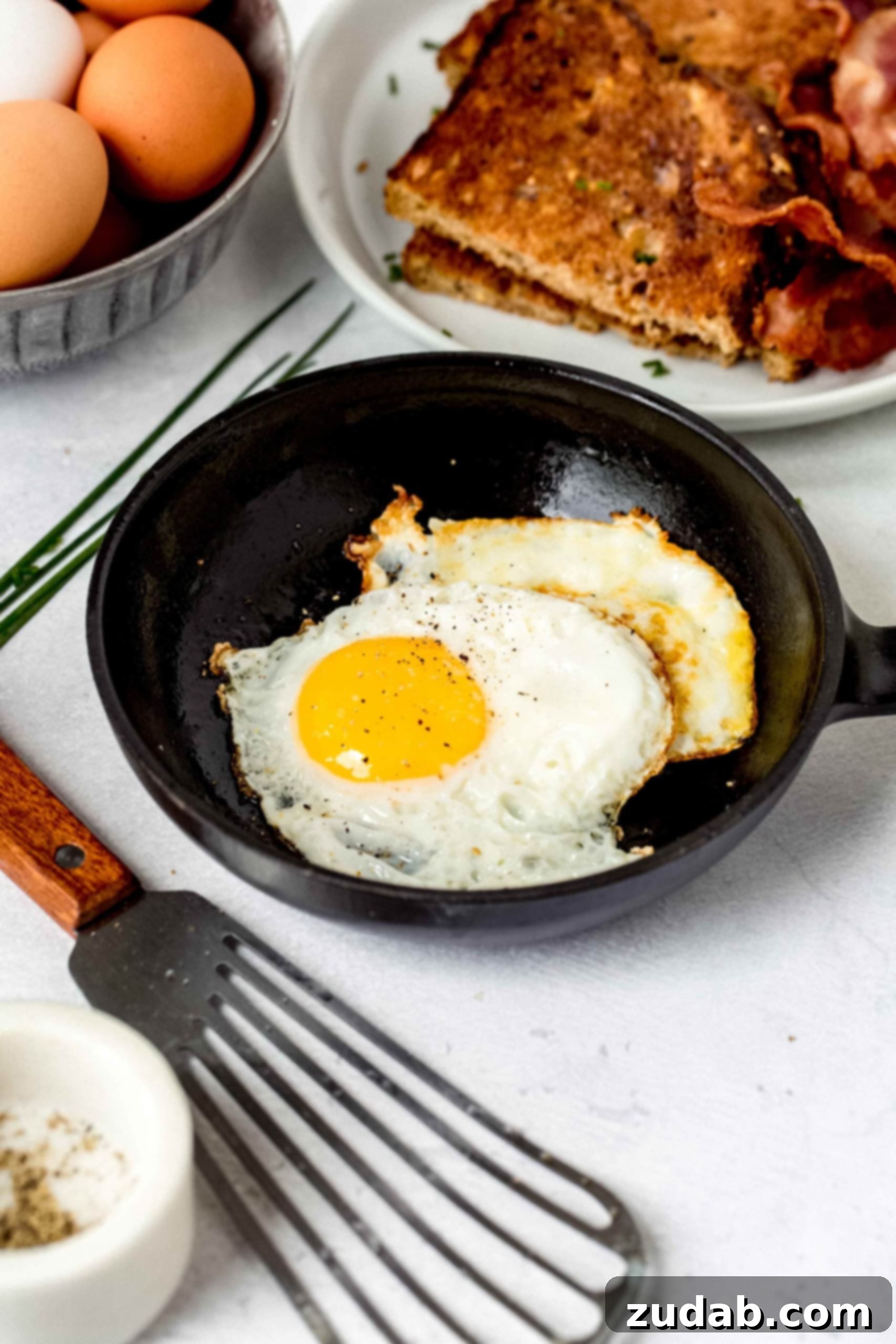 Mastering the Art of the Fried Egg 2 Two perfectly fried eggs with golden yolks and crispy, lacy white edges, resting in a small black frying pan. A flexible spatula is placed in front of the pan, and a plate of golden brown toast along with a carton of fresh brown eggs are visible in the background, hinting at a delightful breakfast.