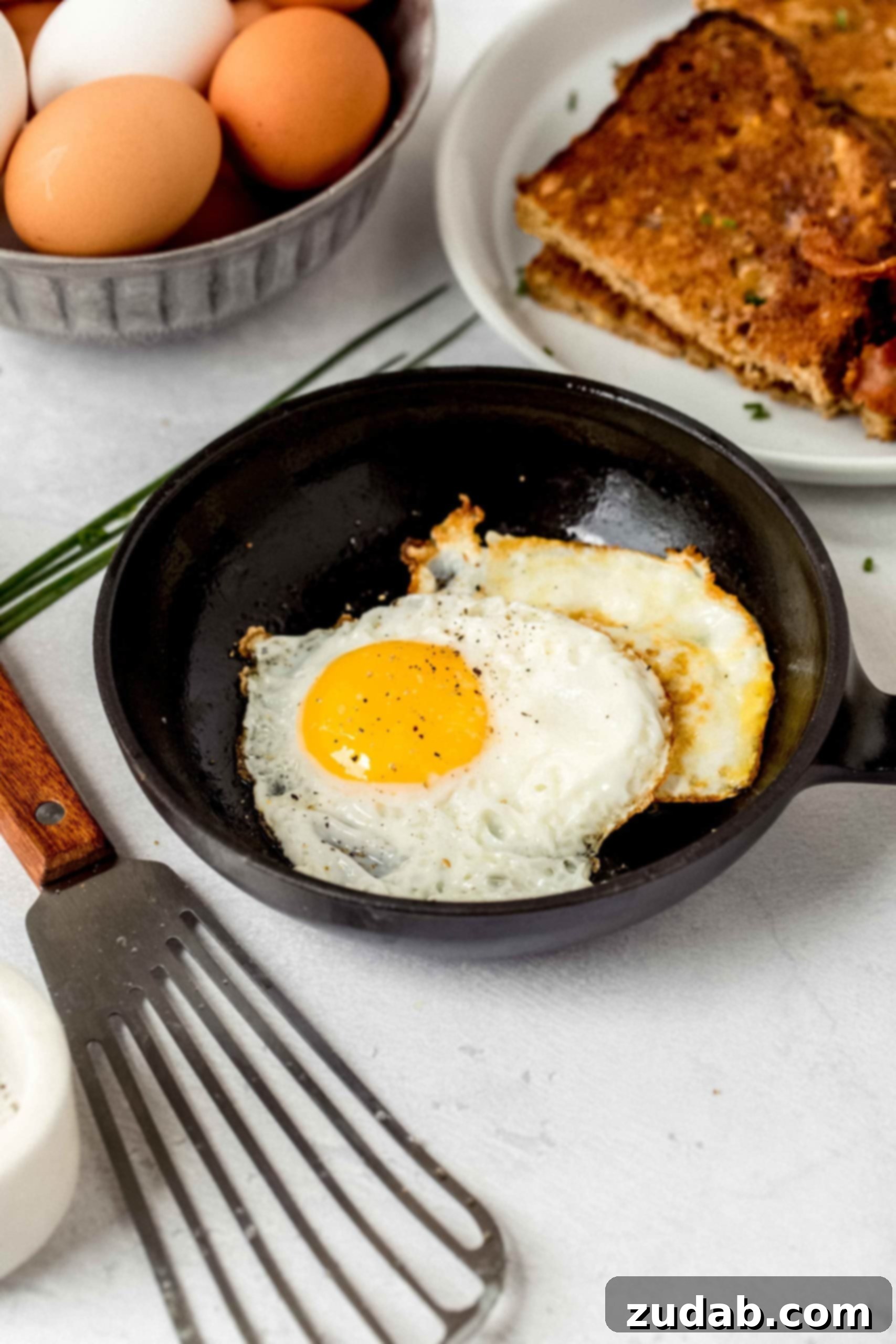 Mastering the Art of the Fried Egg 5 Two golden-fried eggs with bright, runny yolks are cooking in a small black nonstick frying pan. In the background, a carton of fresh eggs and a plate filled with crispy bacon and perfectly toasted bread set the scene for a delicious breakfast. A fish spatula rests on the counter next to the pan, ready for use.