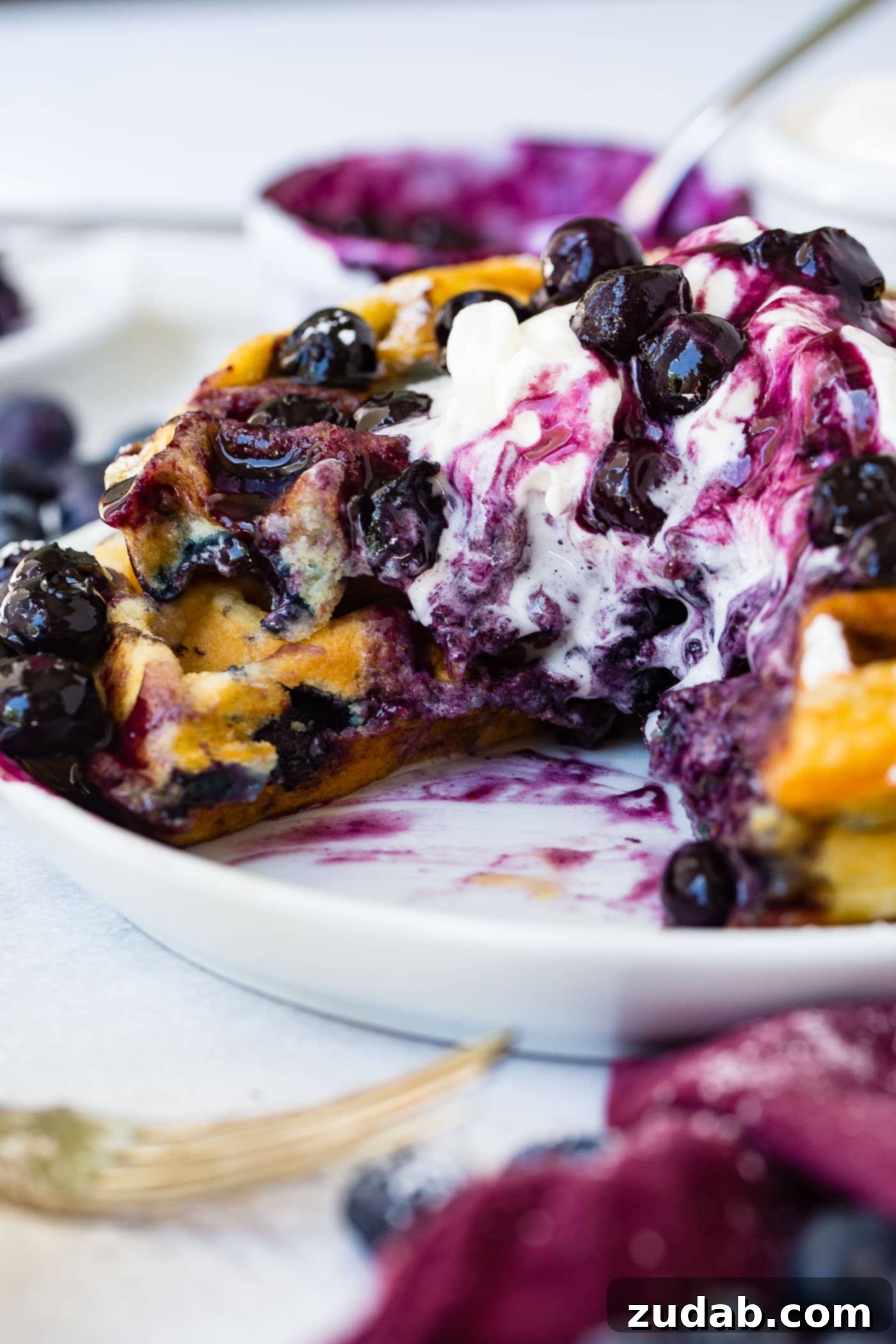 Blueberry Waffles A partially eaten stack of golden blueberry waffles, generously topped with whipped cream and fresh blueberries, highlighting the berries baked inside.