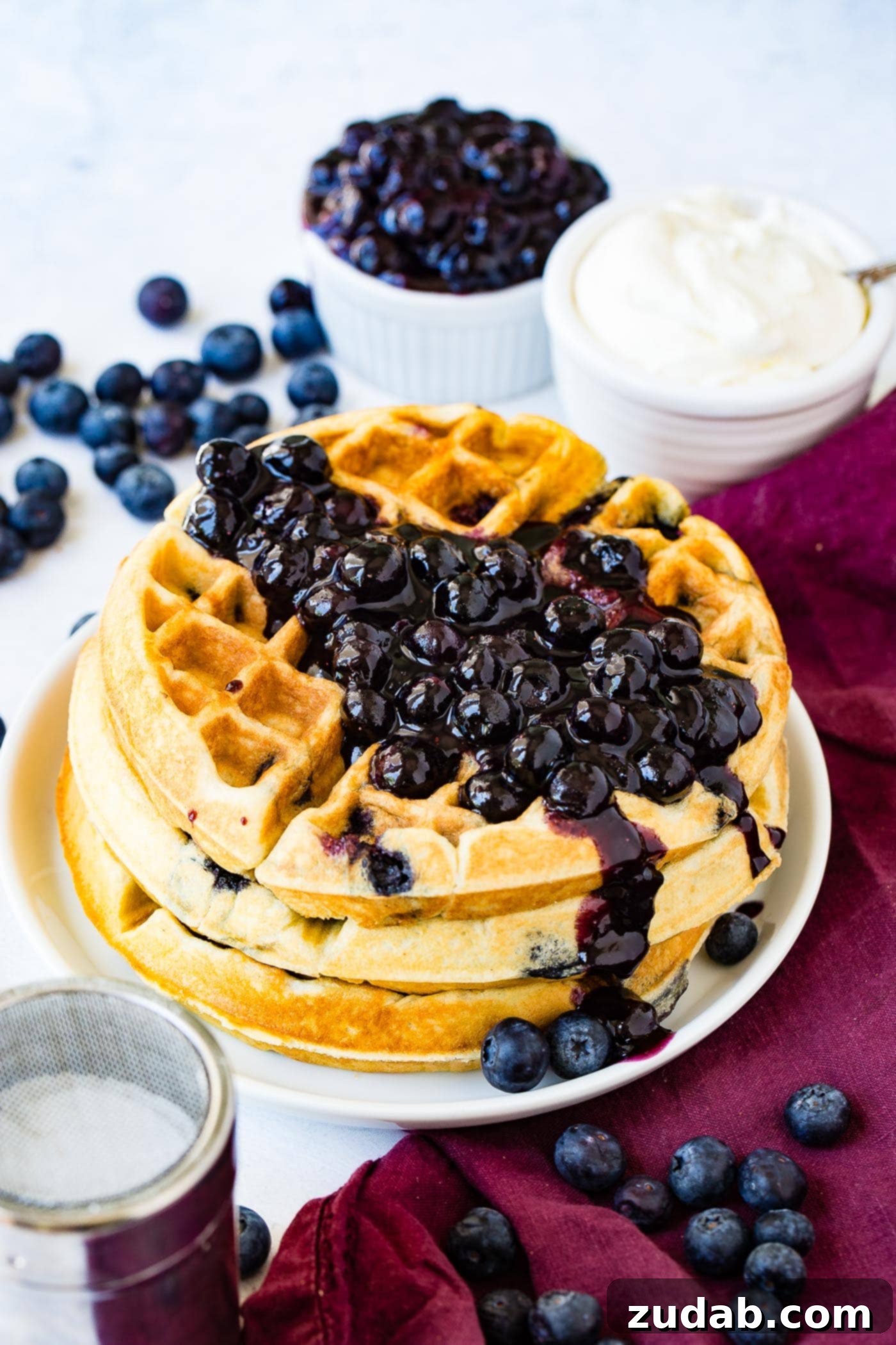 Blueberry Waffles A stack of three fluffy blueberry waffles, generously garnished with fresh blueberries and a dollop of whipped cream, with more berries in the background.