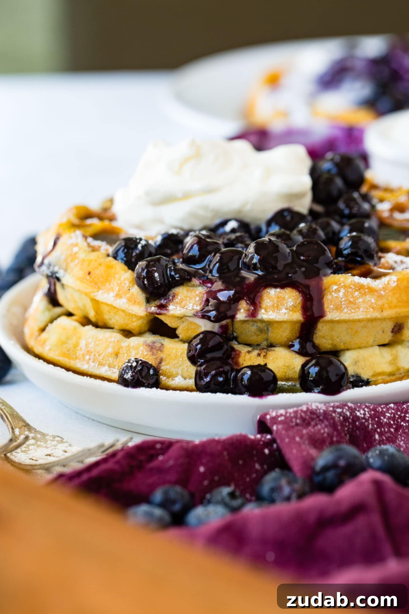 Blueberry Waffles A stack of golden blueberry waffles on a dinner plate, generously covered with fresh blueberries and a mound of whipped cream, with more berries on the side.