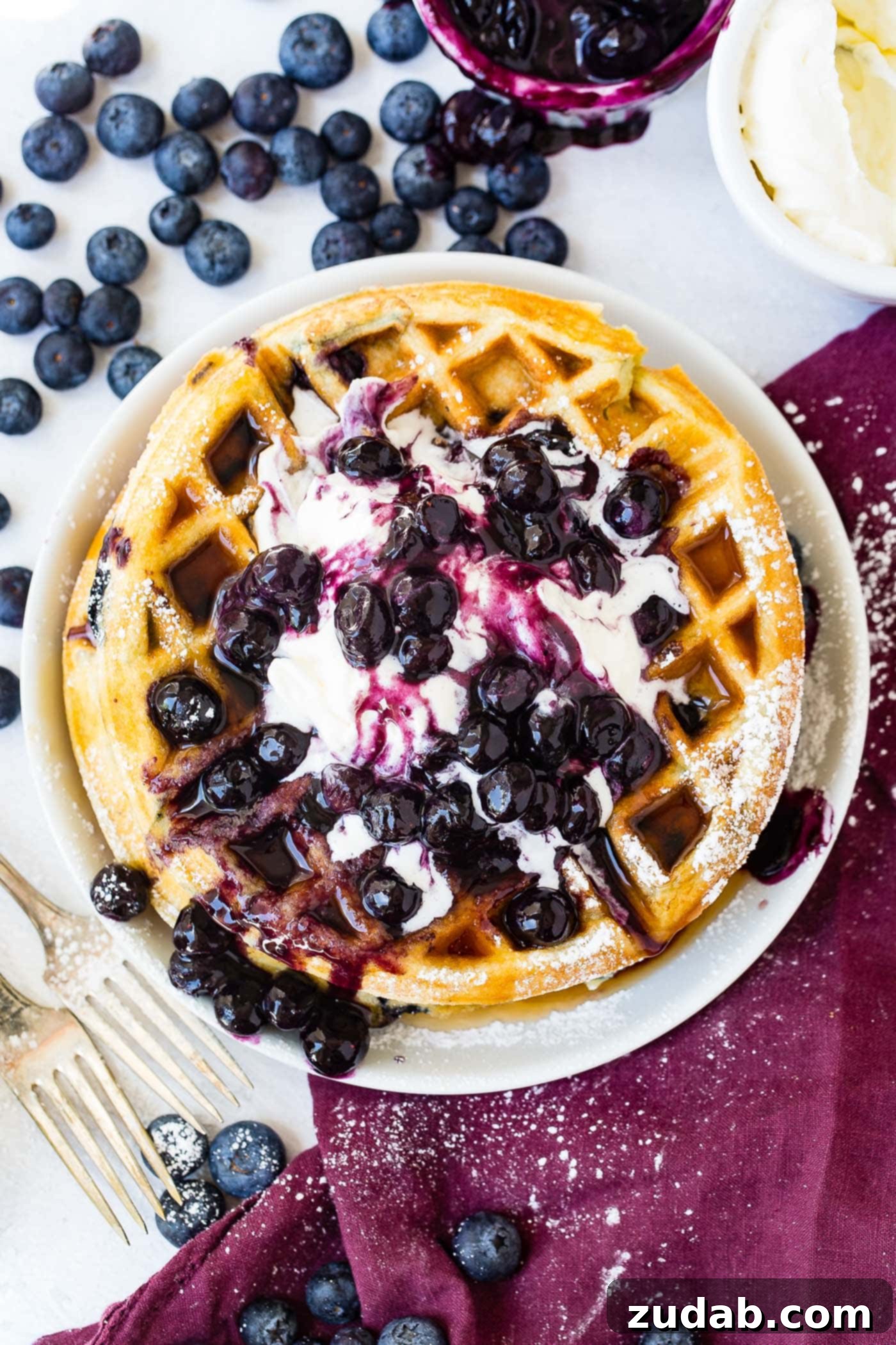 Blueberry Waffles A top-down view of a golden blueberry waffle, adorned with whipped cream and fresh blueberries. In the soft-focus background are more blueberries, a bowl of mashed blueberries, and a bowl of whipped cream, all on a wine-colored napkin.